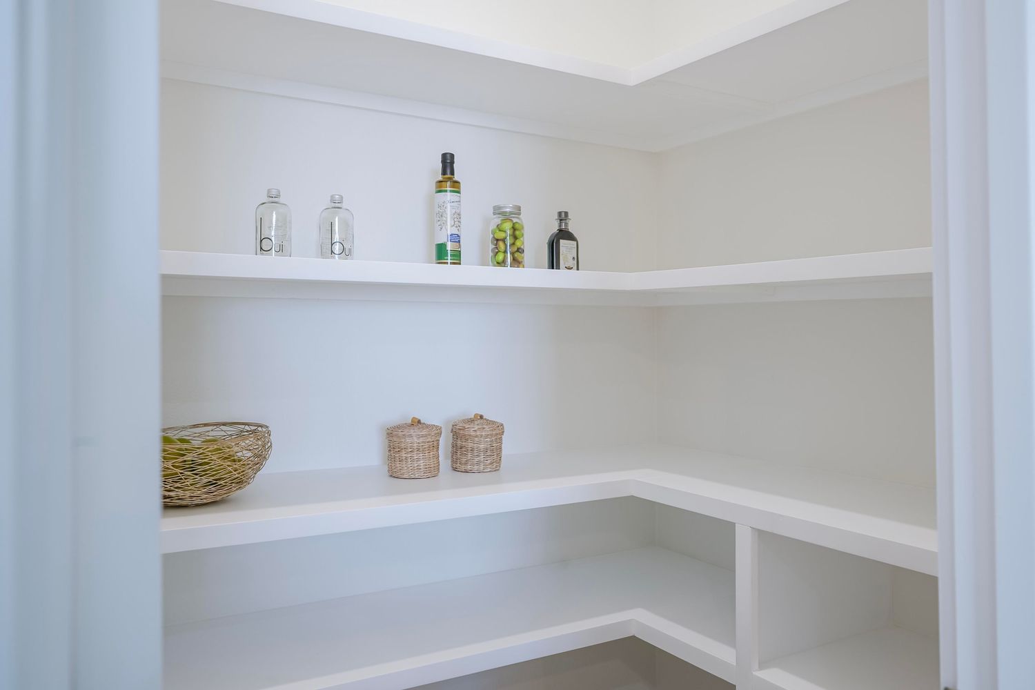 White corner pantry with built-in shelving displaying glass bottles and storage baskets.
