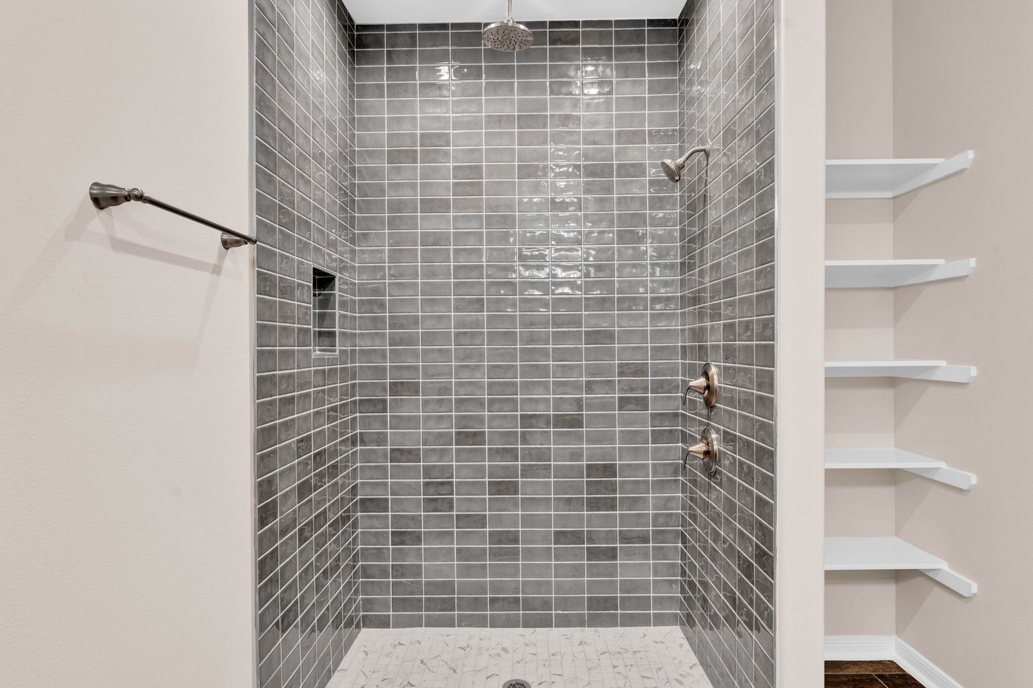 Modern walk-in shower with gray subway tiles, rose gold fixtures, pebble floor, and built-in white shelving.