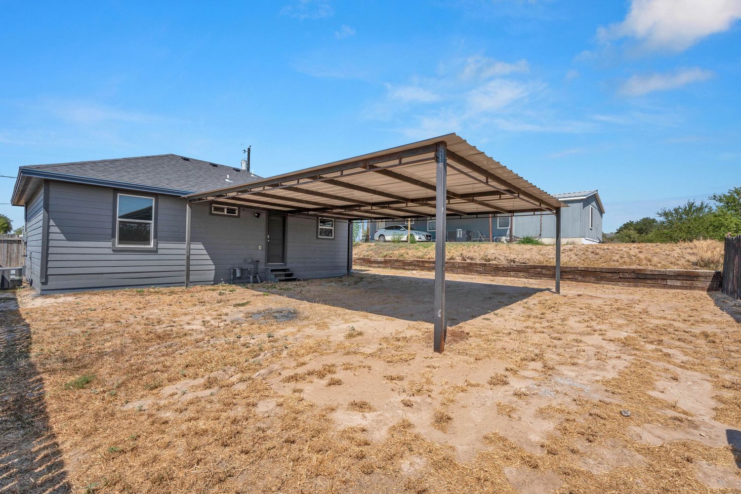 Gray ranch home with large metal patio cover over dry yard, blue sky backdrop.