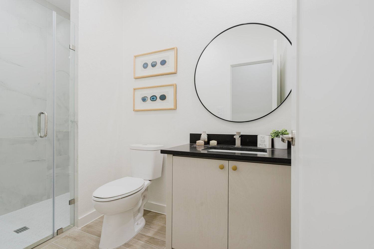 Elegant bathroom featuring glass shower, beige vanity, and circular mirror with wall art.