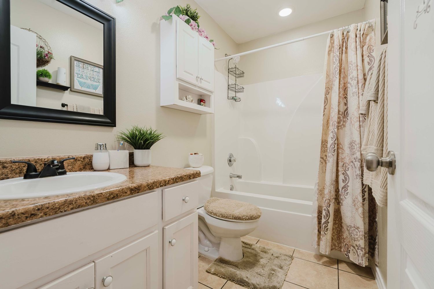 A modern white bathroom featuring granite countertops, white cabinets, and a shower with patterned curtain and decorative plants.