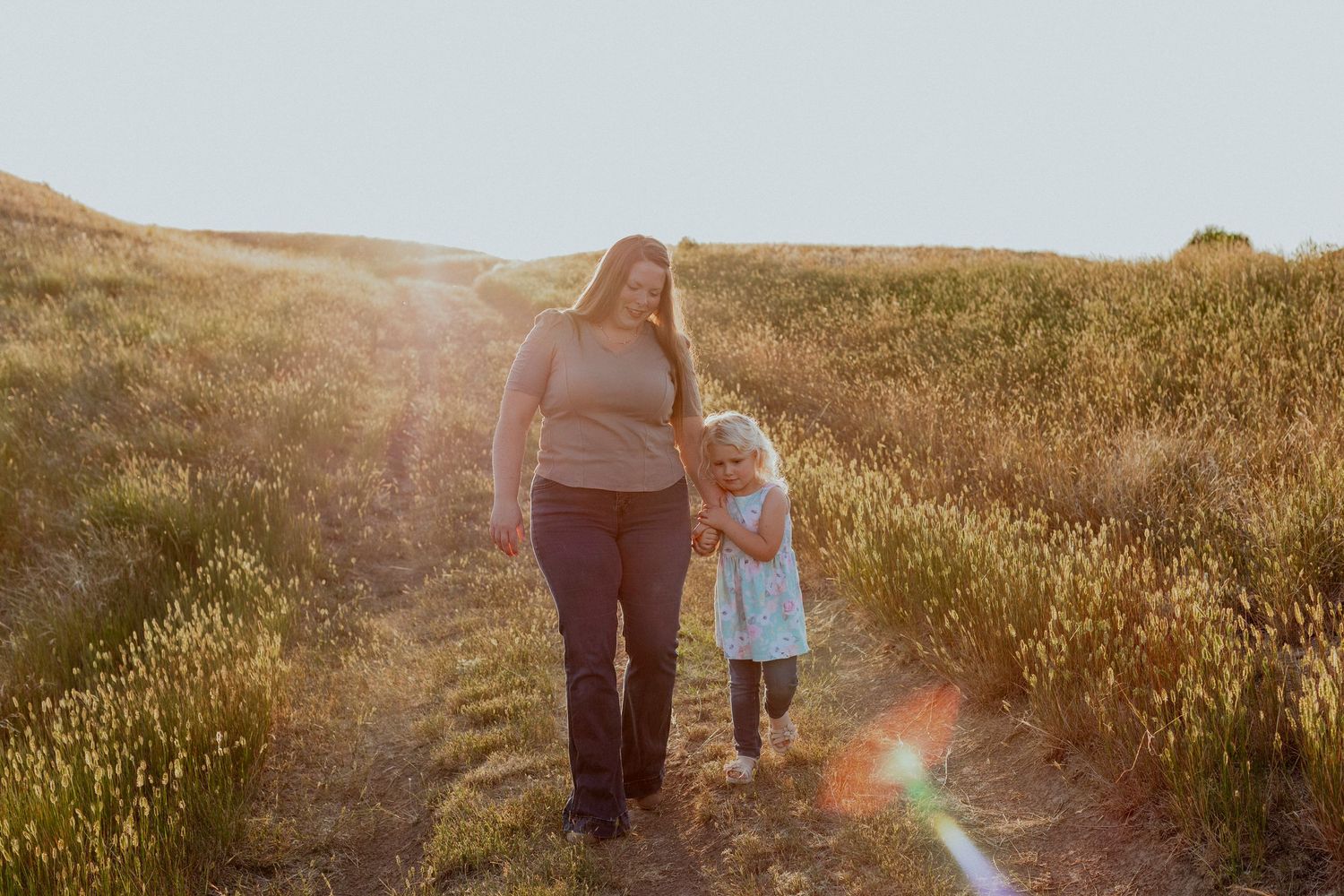 Two figures walk hand in hand along a sunlit path through golden fields at sunset.
