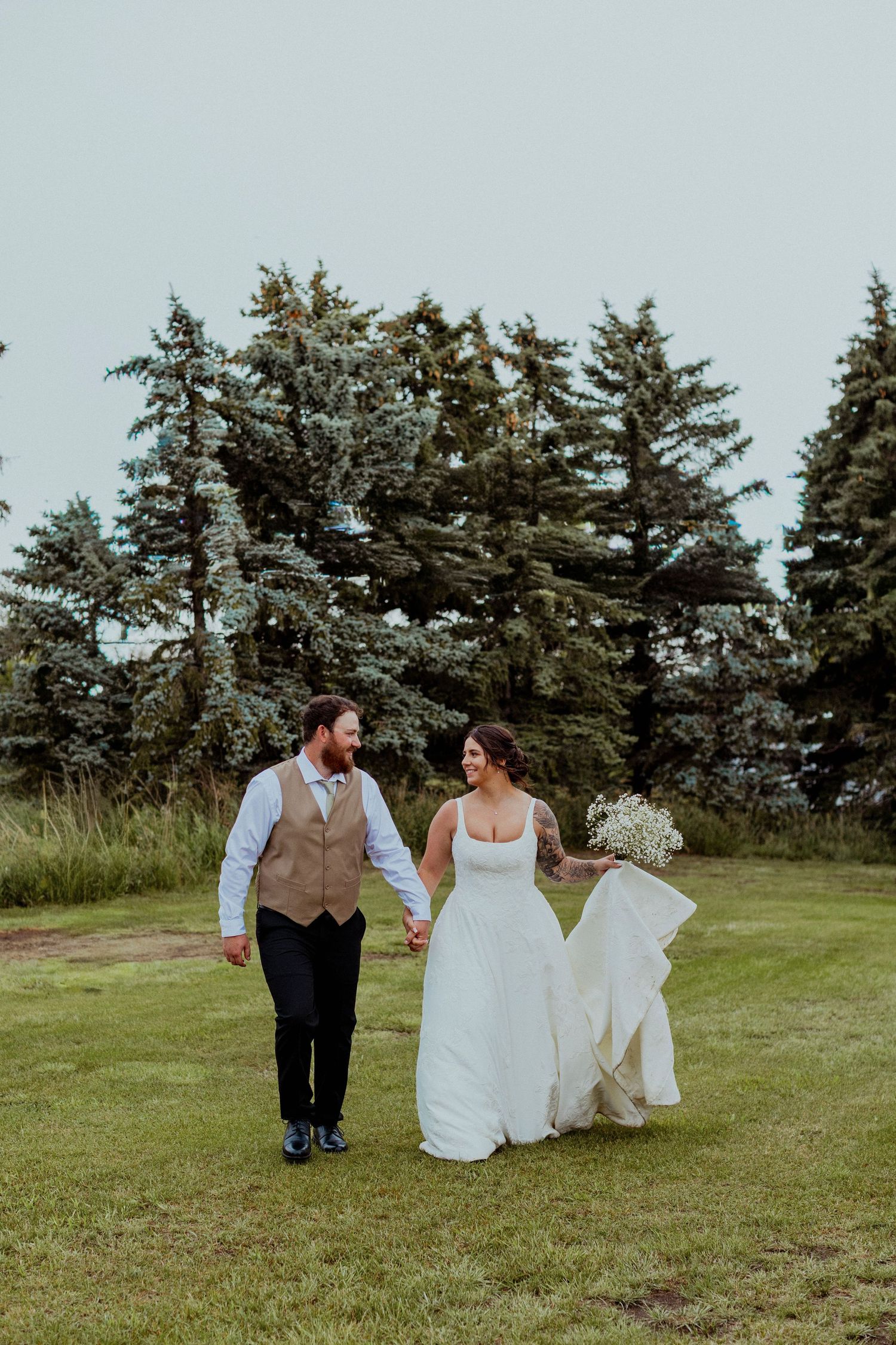 A couple in wedding attire walks hand in hand through a grassy field with evergreen trees in the background.