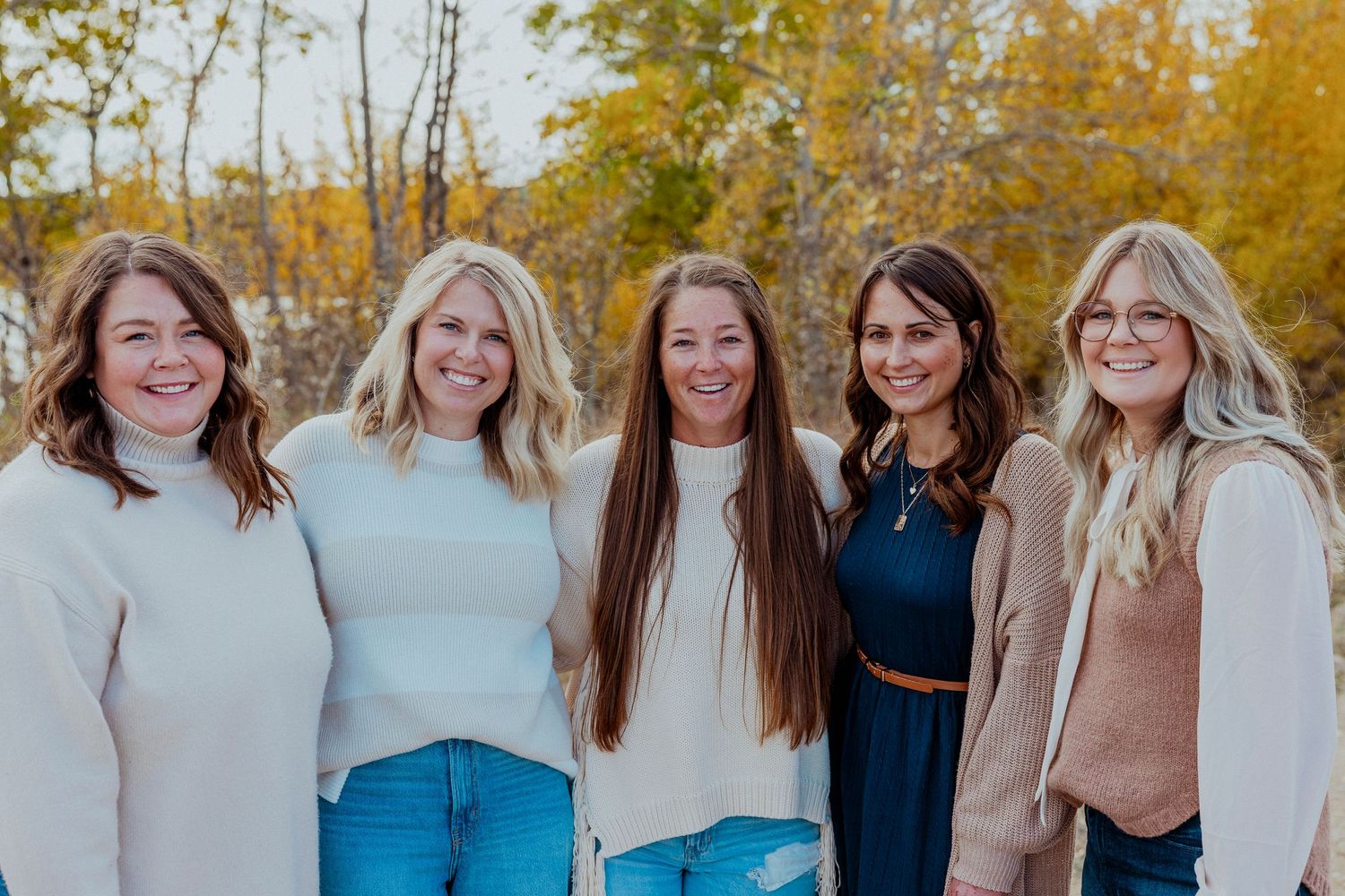 A group of people stand close together smiling outdoors during autumn with fall foliage in the background.