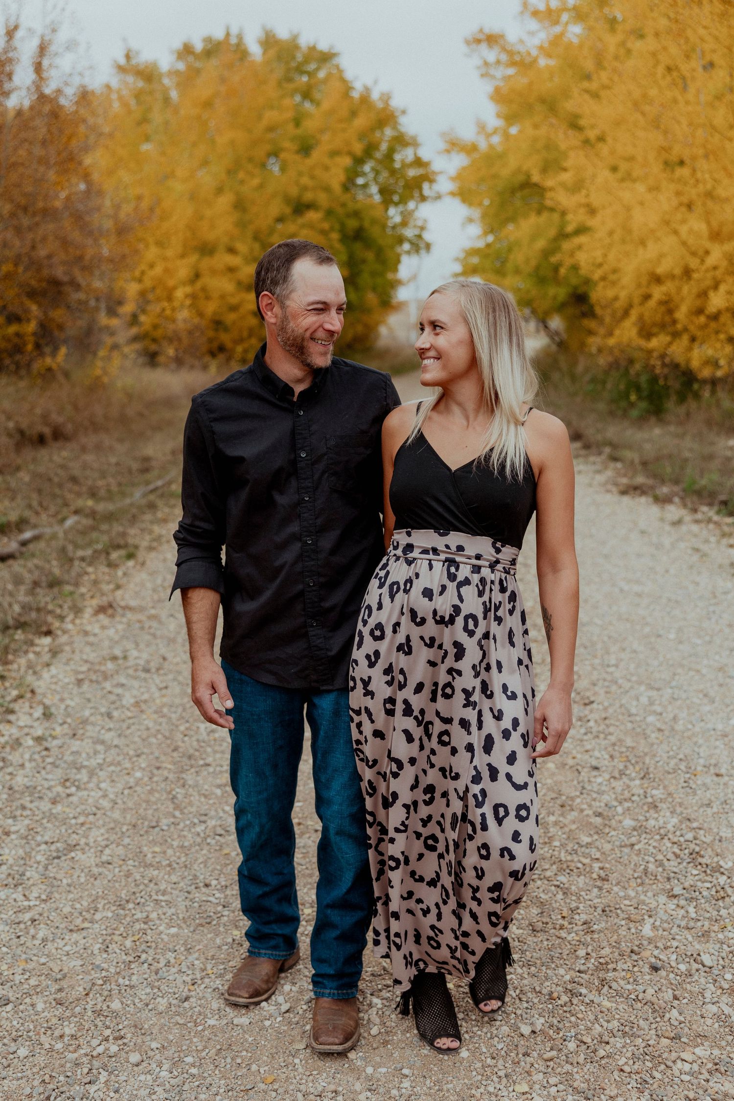 A couple walks together on a gravel path surrounded by golden autumn trees in the background.