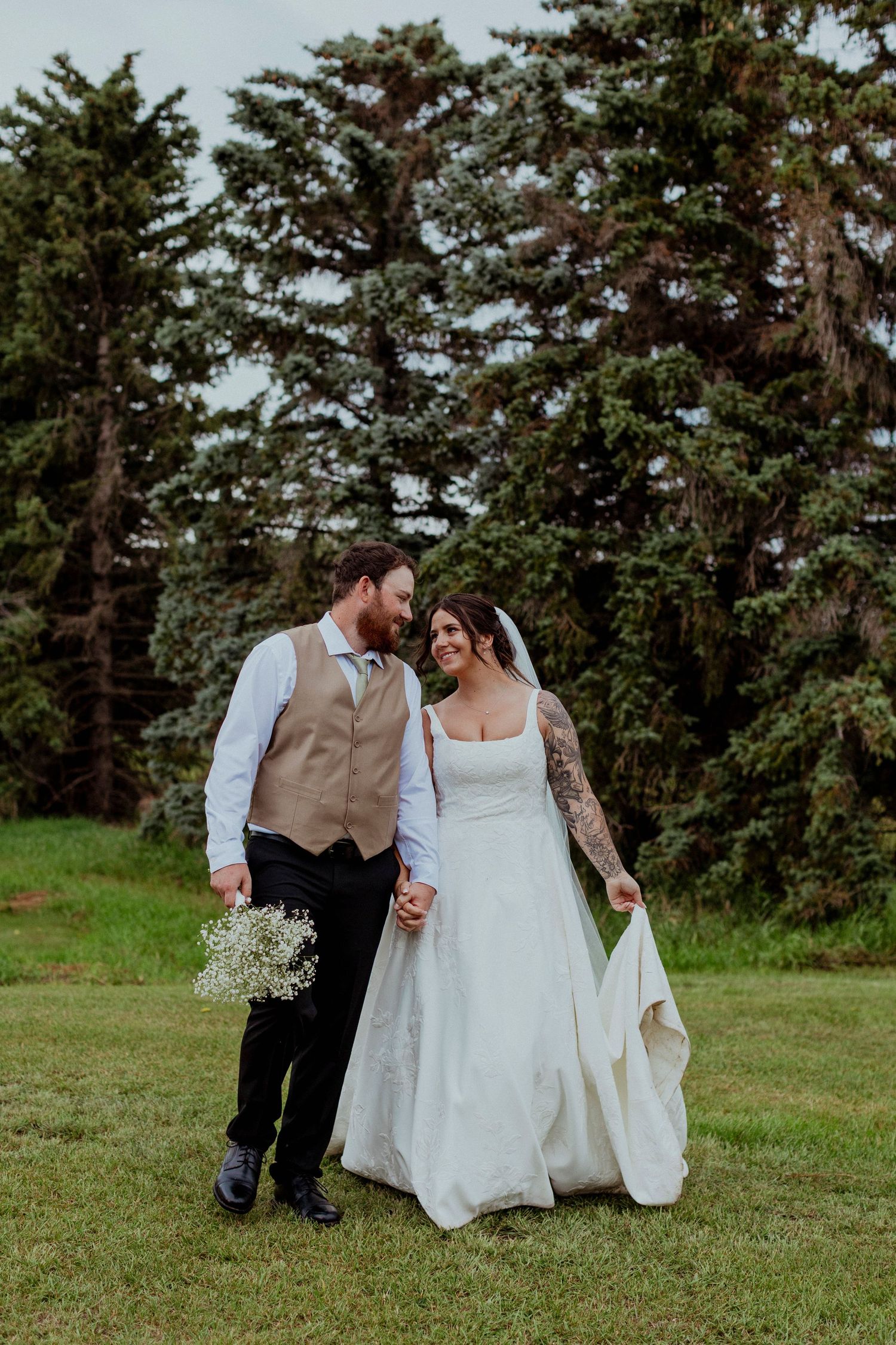 A couple sits together on green grass surrounded by tall evergreen trees during their outdoor forest wedding.