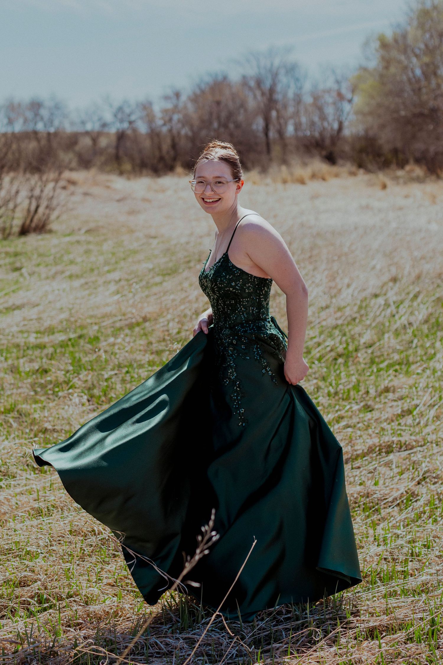An emerald green formal gown flows gracefully in the breeze against a rustic outdoor landscape on a sunny day.