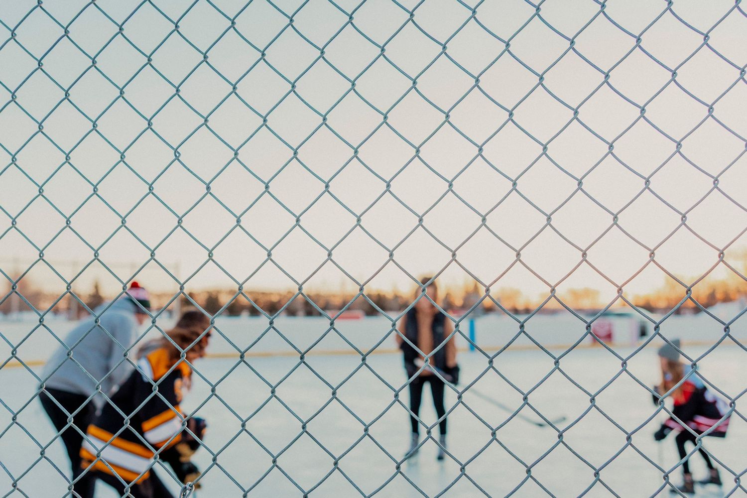 A chain link fence stretches across a snowy field with blurred figures in the distance.