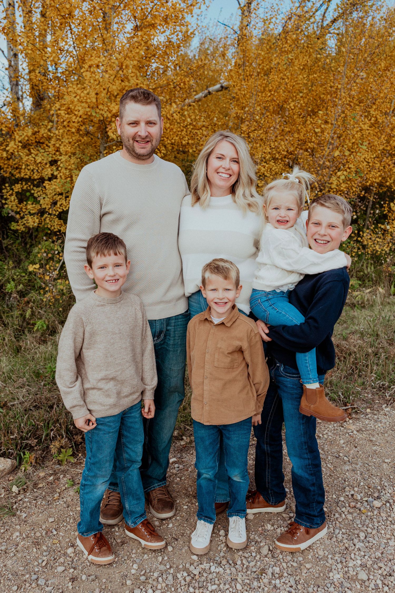 A family wearing coordinated neutral outfits poses together against a backdrop of golden autumn trees.