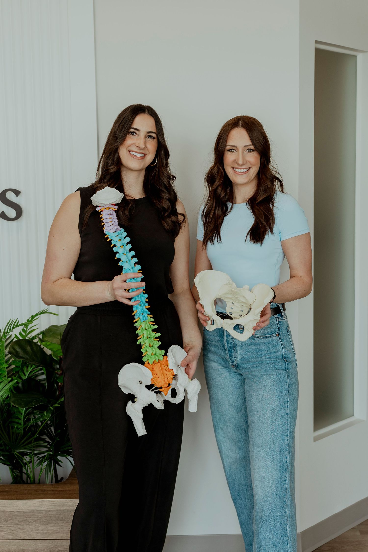 Two healthcare professionals in a medical office holding anatomical models of a spine and pelvis. Chiropractor content, social media, sask