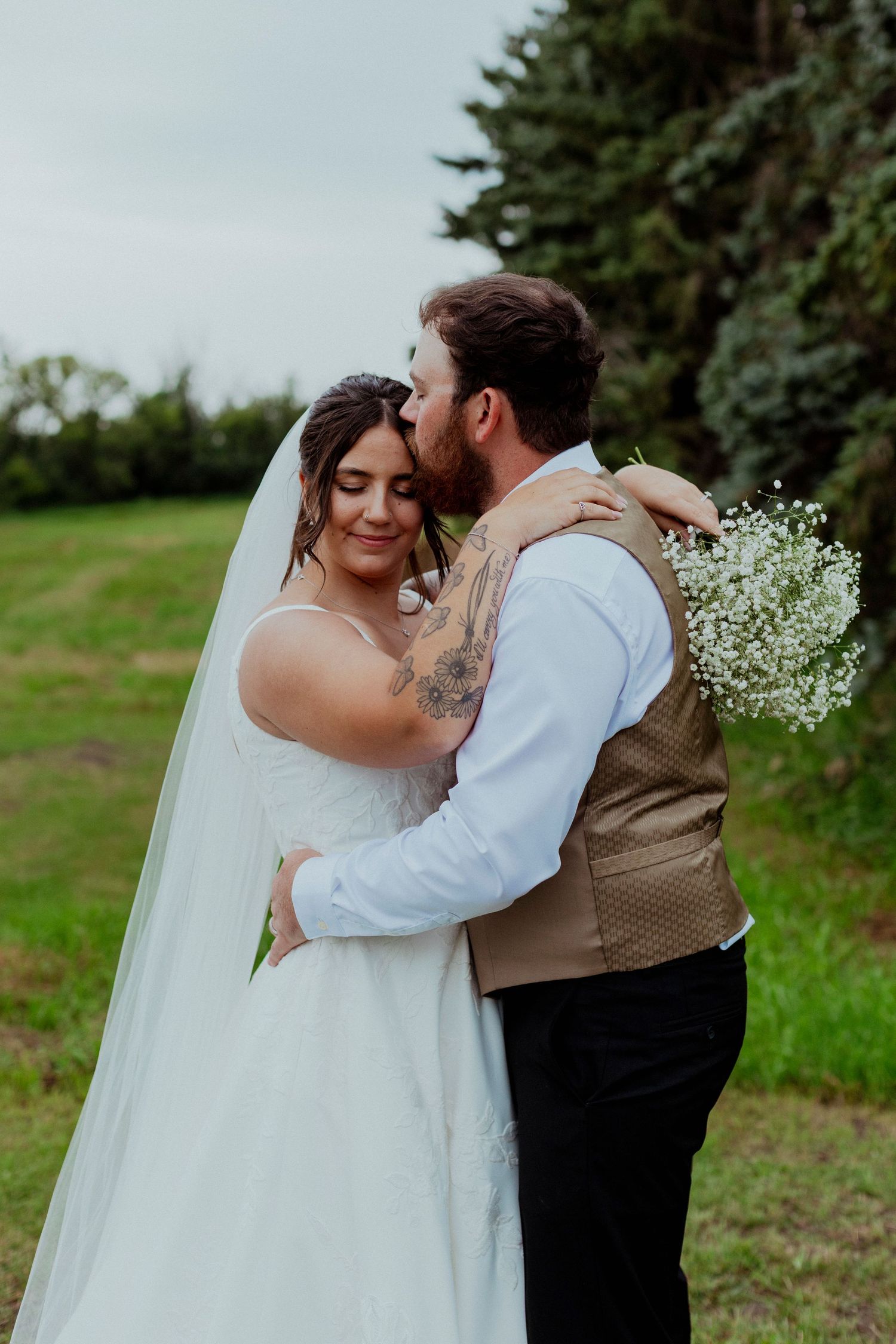 A romantic newlywed couple embraces in a lush green outdoor setting with the bride wearing a flowing white dress.