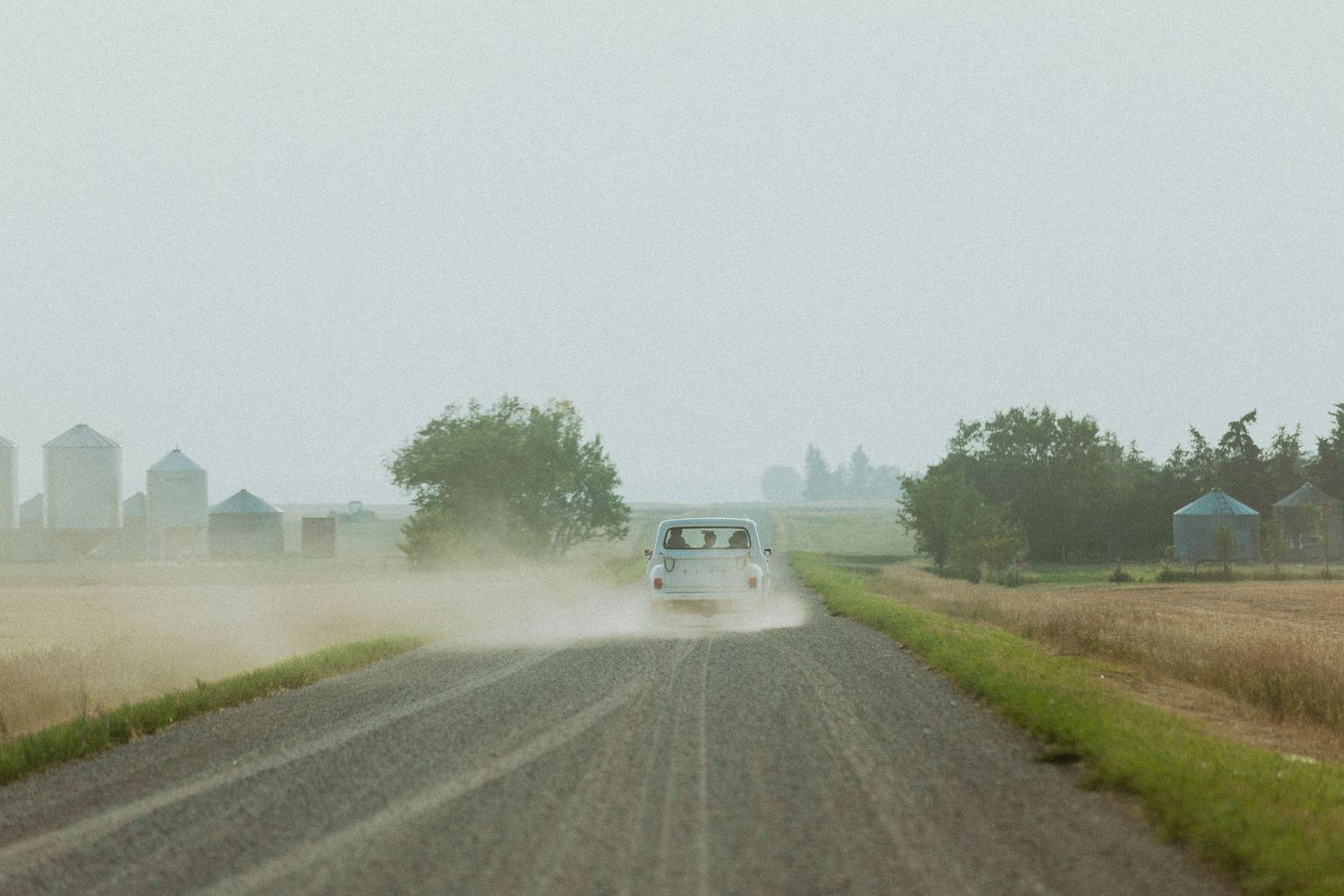 Car drives down dusty rural road on misty day with grain silos visible in background.