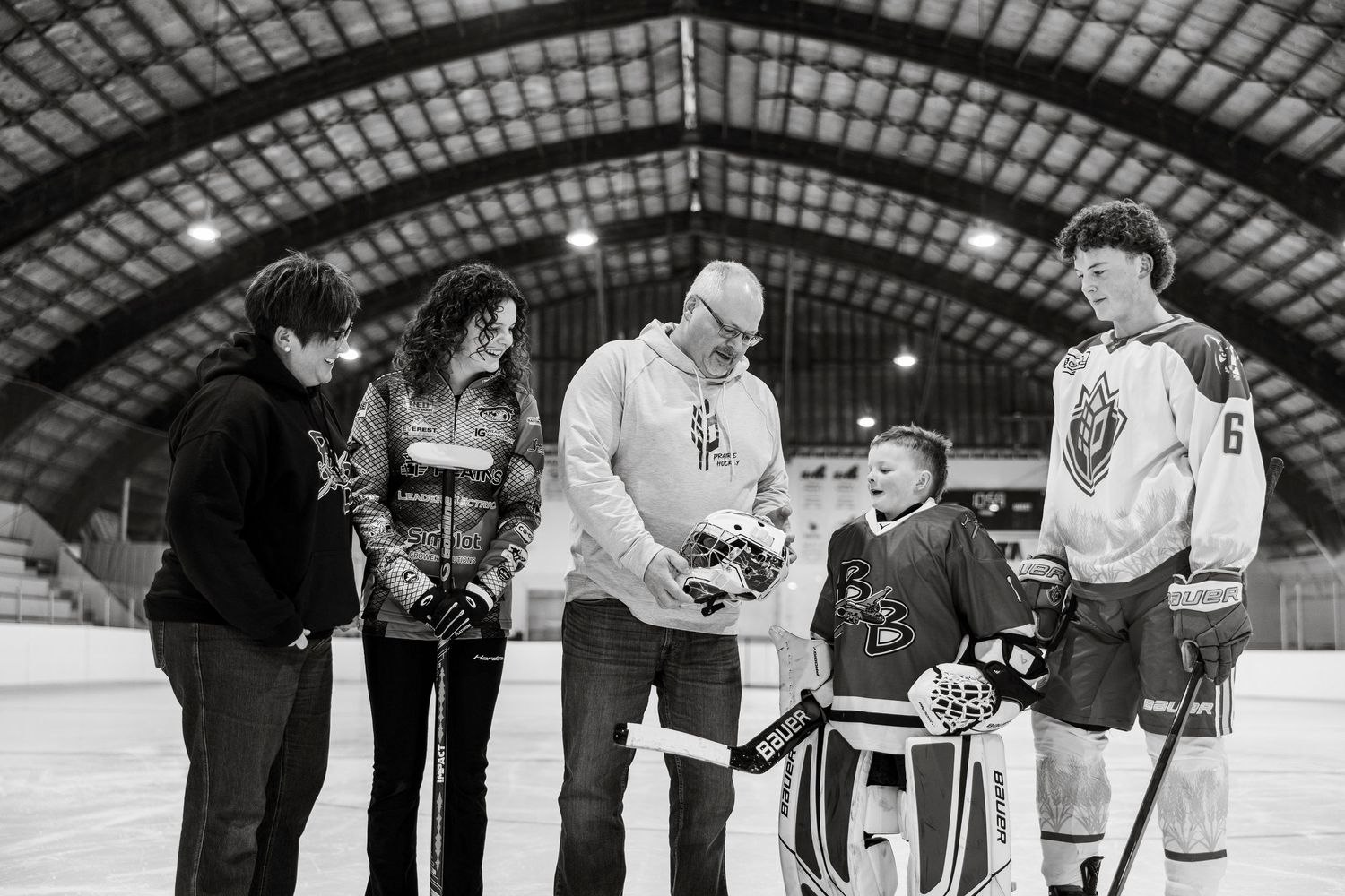 Hockey players gather on an ice rink during practice session in an indoor arena with metal roof structure.
