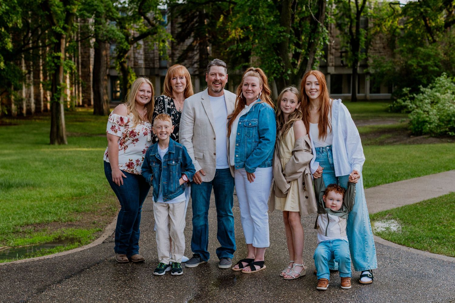 A group of people in coordinated blue and beige outfits pose together on a tree-lined path in a park setting.