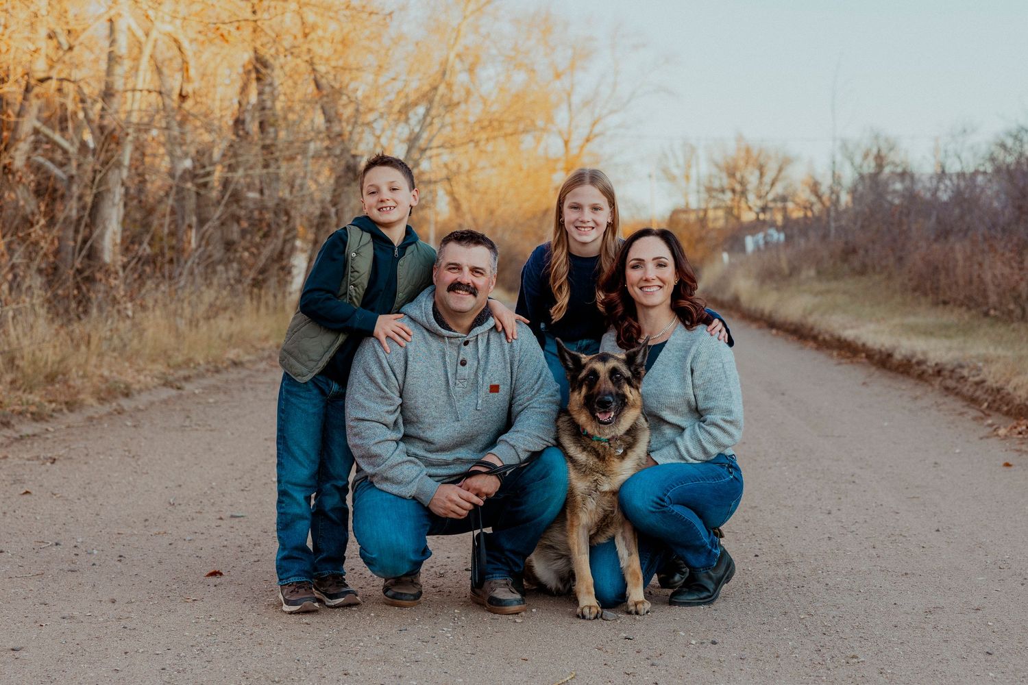 A group of people and their German Shepherd dog pose together on a dirt path during autumn.