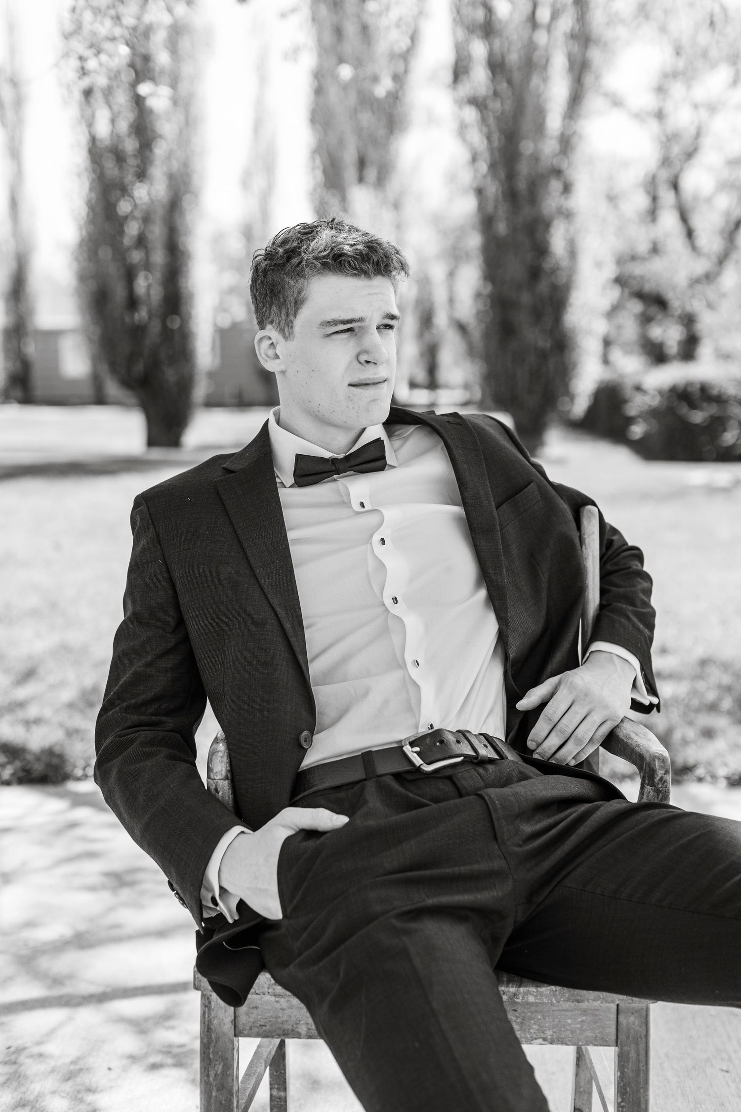 Black and white portrait of person in formal attire with bow tie sitting on chair outdoors in a park-like setting. graduate, sask grad