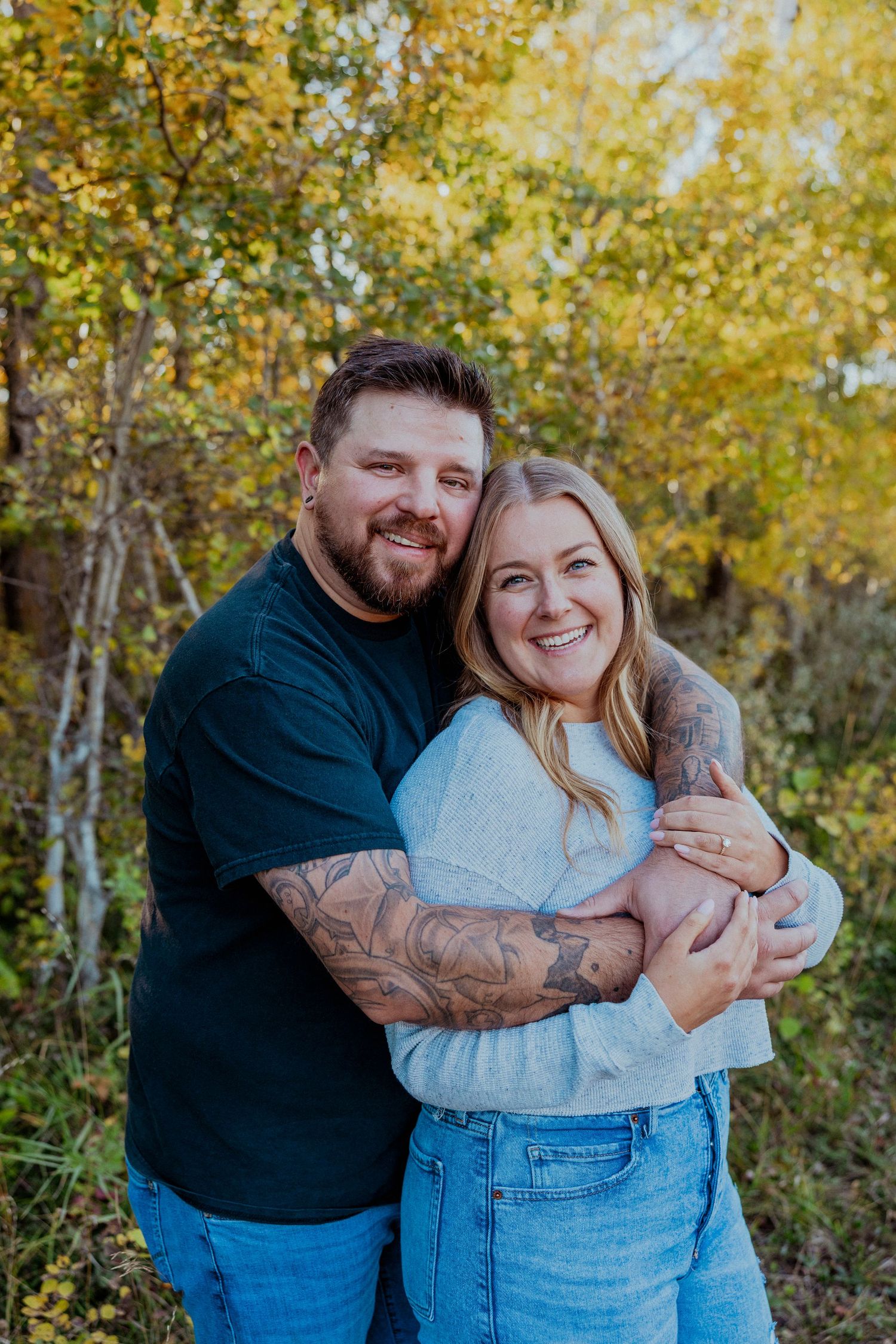 Couple embraces in autumn forest setting with golden leaves and sunlight filtering through trees.