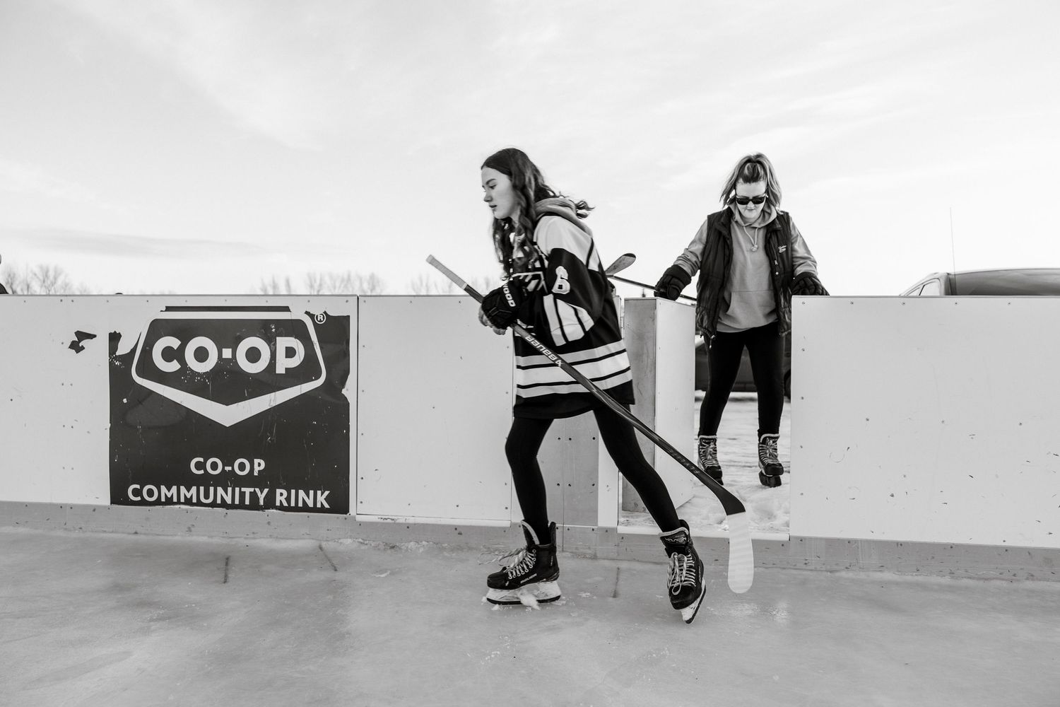 female hockey players enter an outdoor rink with CO-OP signs visible in the background. rink photography session life capture winter in sask