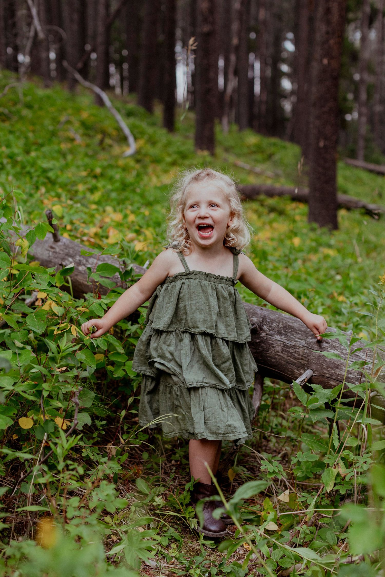 Young child in green dress running joyfully through a forest setting.