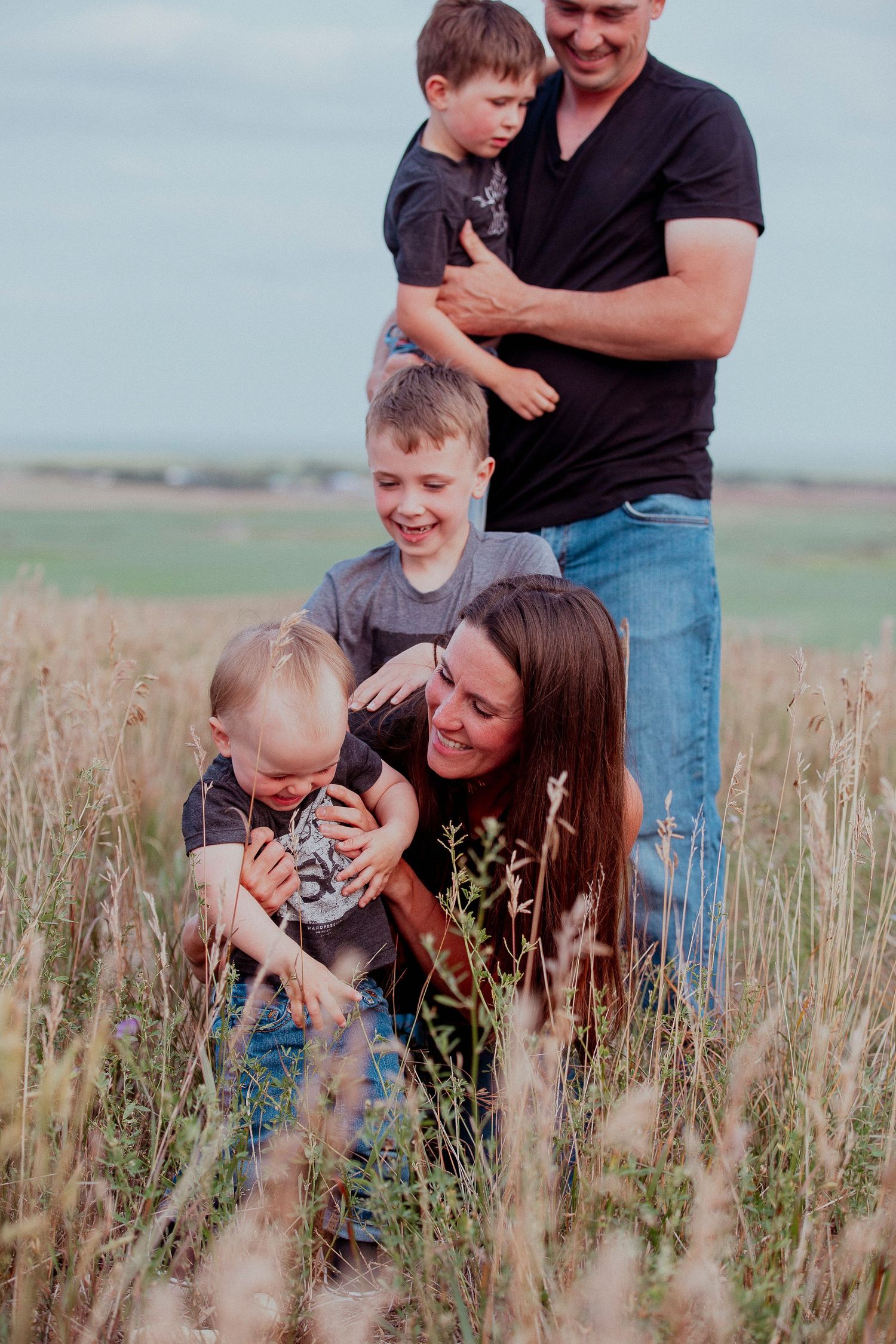 A joyful family shares laughter and embraces while standing together in a field of tall grass at golden hour.