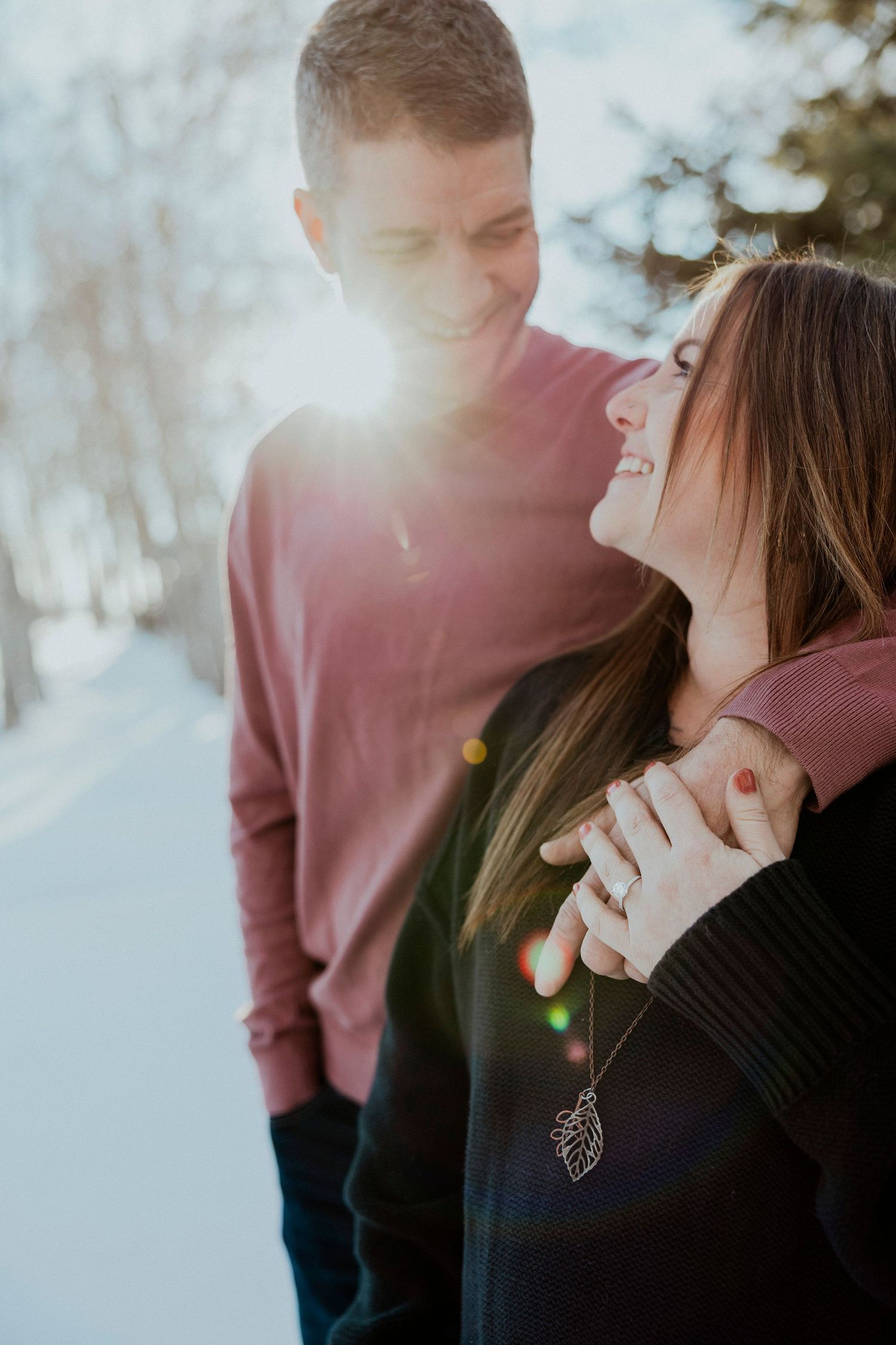 A romantic winter couple shares a tender moment in sunlit snow with sunflare and warm burgundy sweaters.