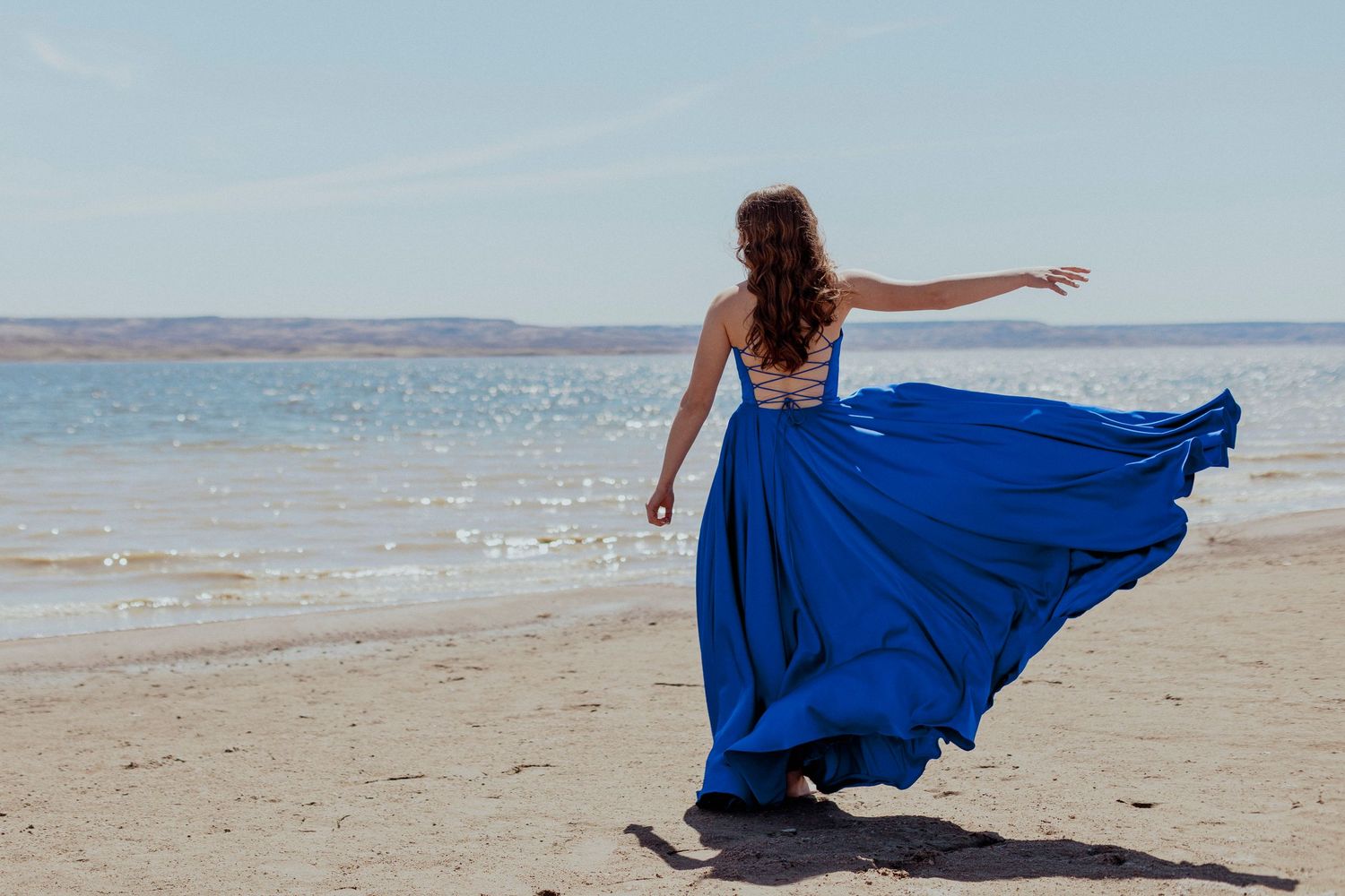 Woman wearing flowing royal blue skirt twirls gracefully on sandy beach with ocean waves and coastline in background.