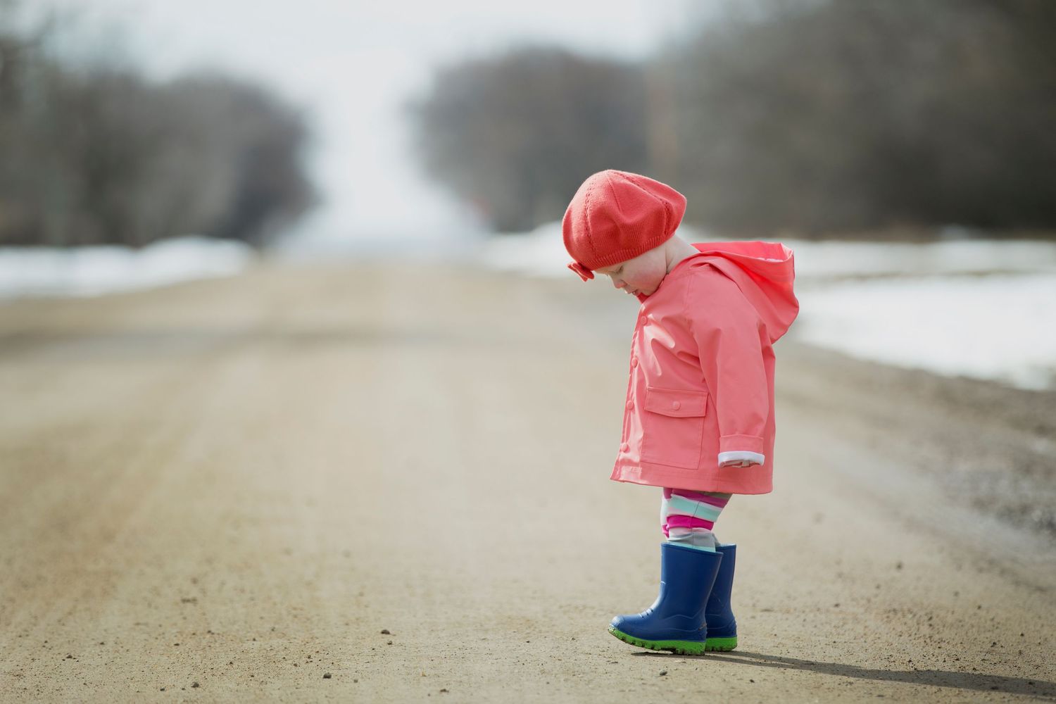 A small figure in a pink winter coat and hat walks away on a snowy dirt path on a foggy winter day.