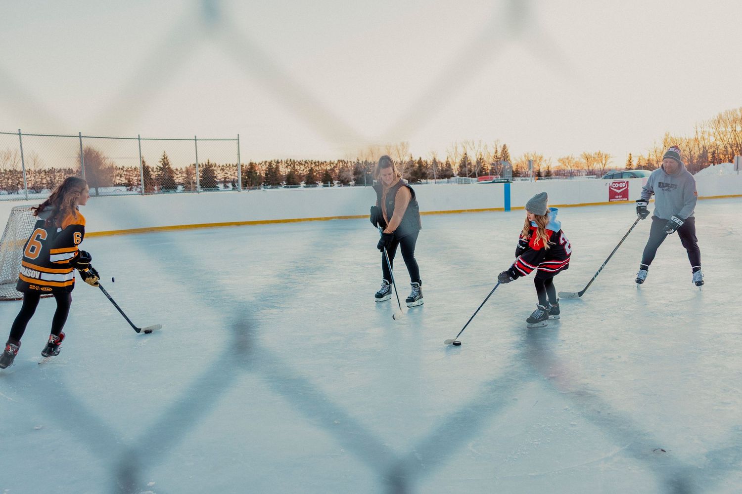 Hockey players skating on an outdoor ice rink during a winter game with snow-covered trees in the background.