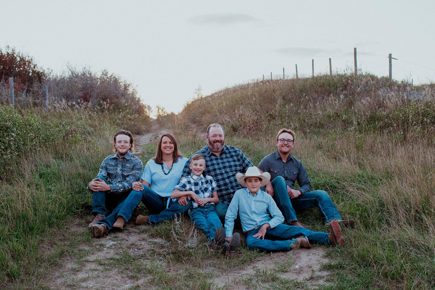 A group sits together outdoors on a grassy hillside at dusk wearing casual clothing and smiling at the camera.