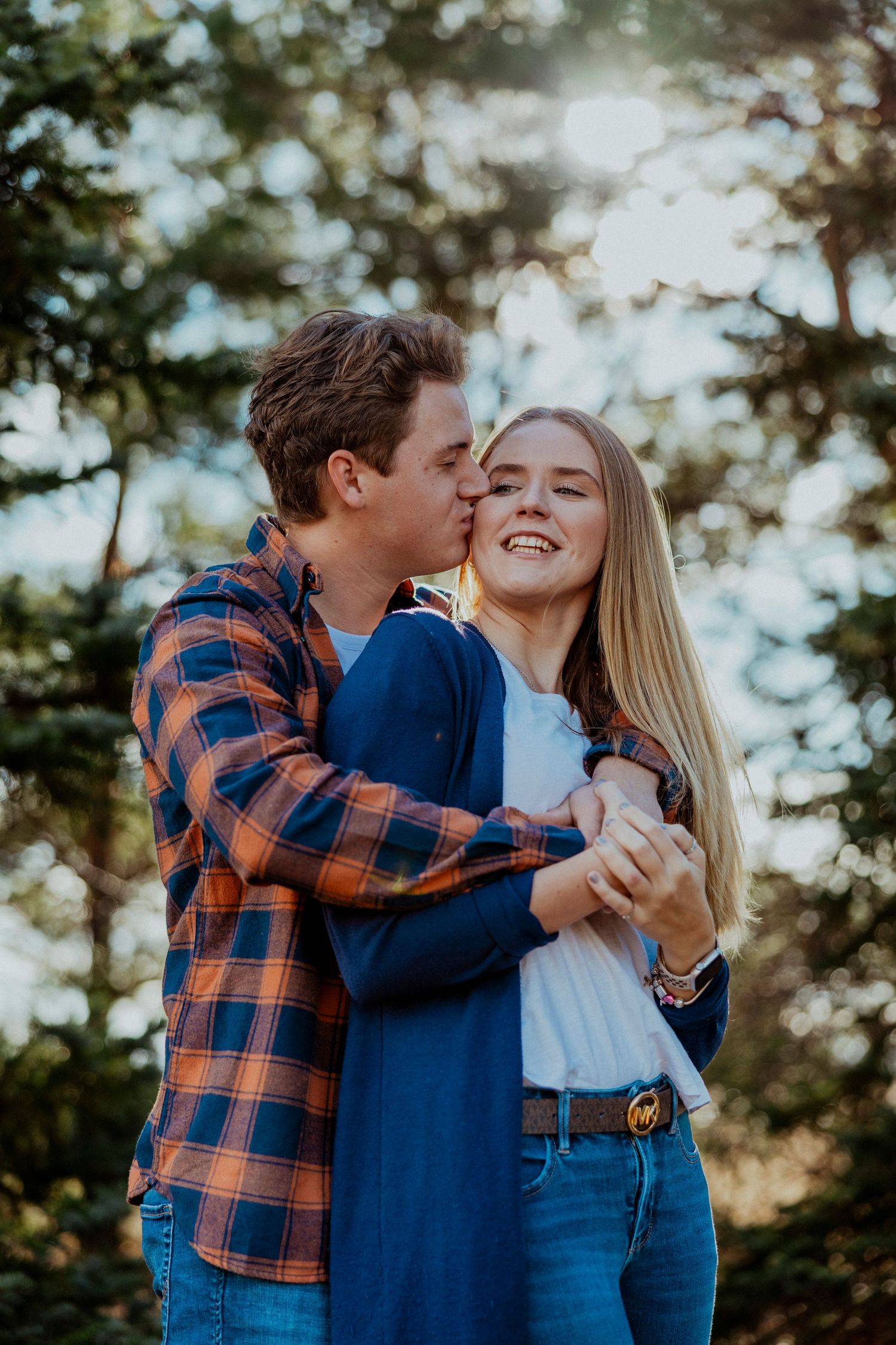 A couple embraces and laughs together outdoors during a romantic photoshoot in a sunlit forest setting.