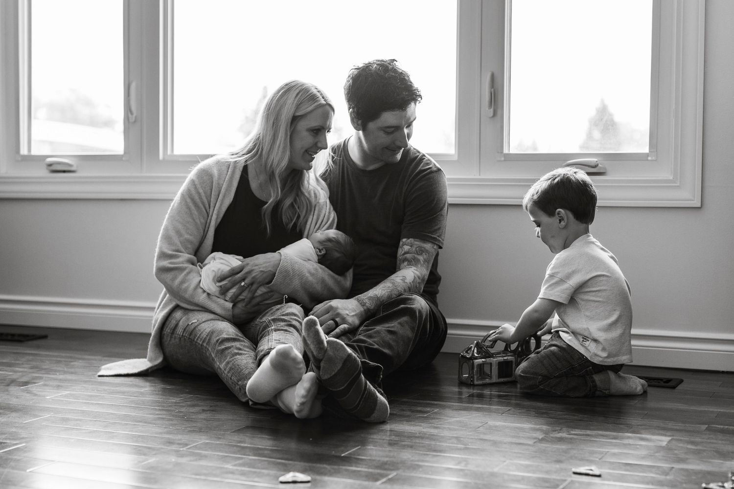 A family sits together on a hardwood floor in a sunlit room during a casual moment at home.