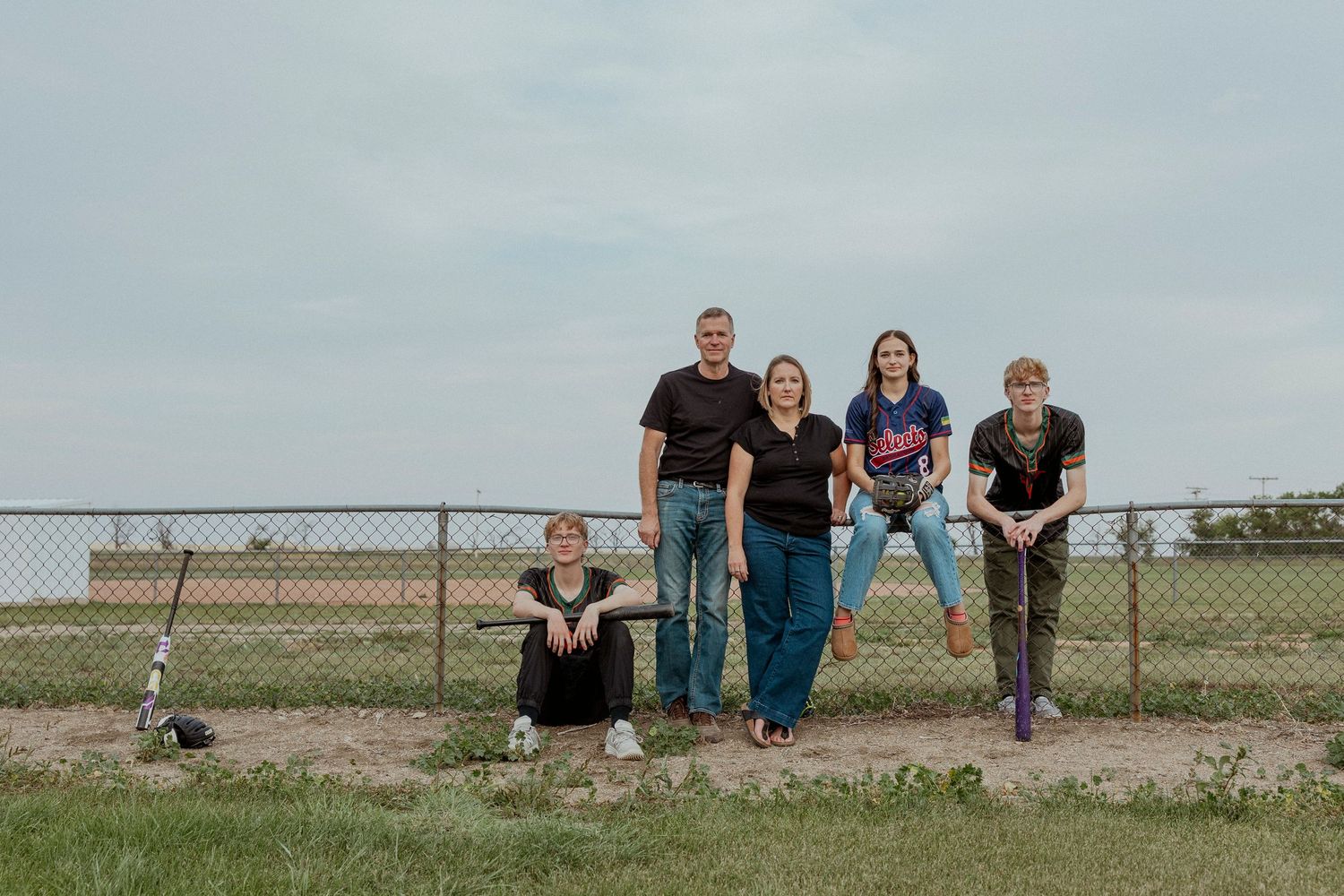 Family of five posing outdoors near a chain-link fence on a cloudy day in a rural setting.