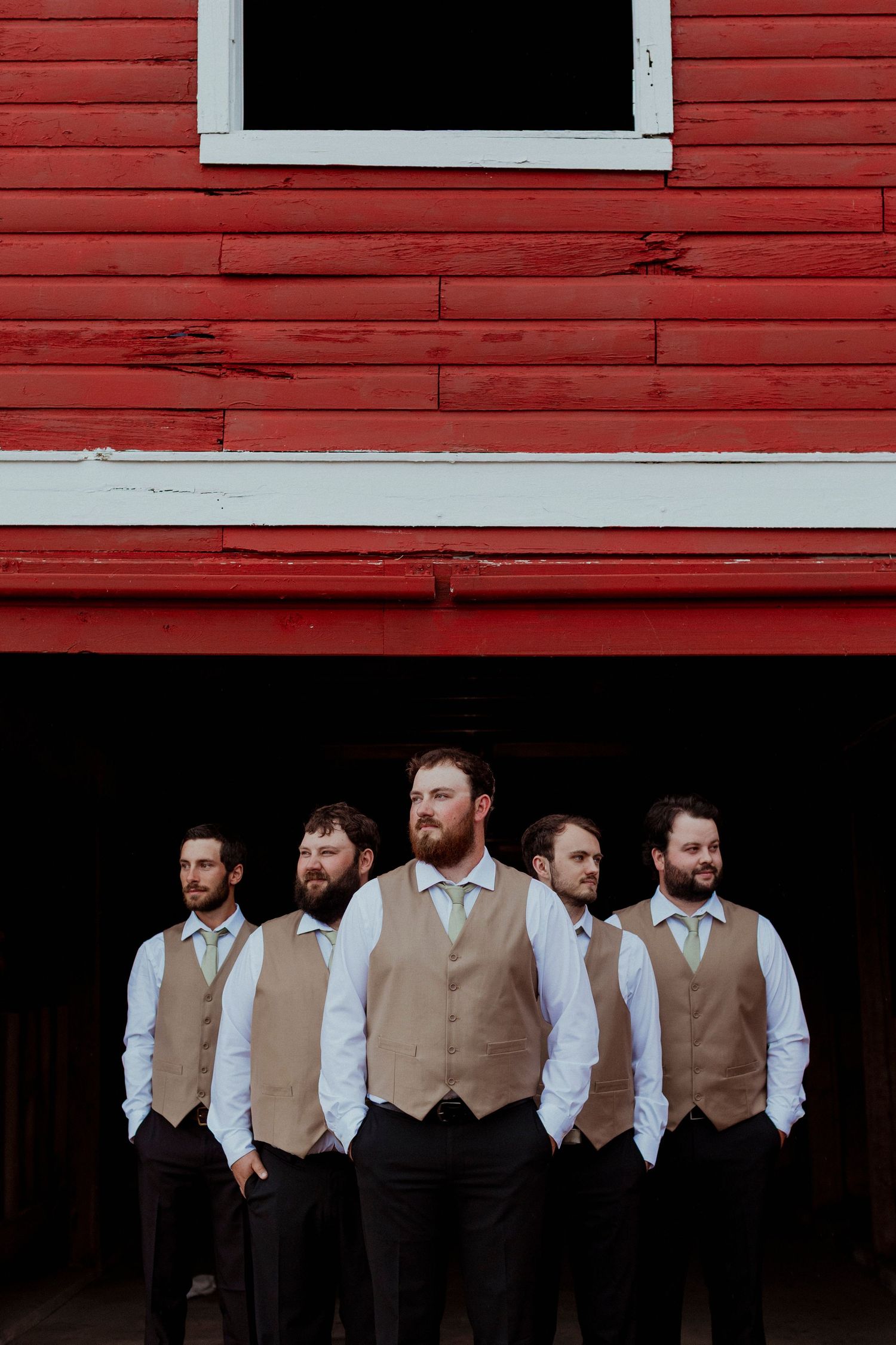 A group of groomsmen in tan vests and white shirts pose in front of a red barn wall.