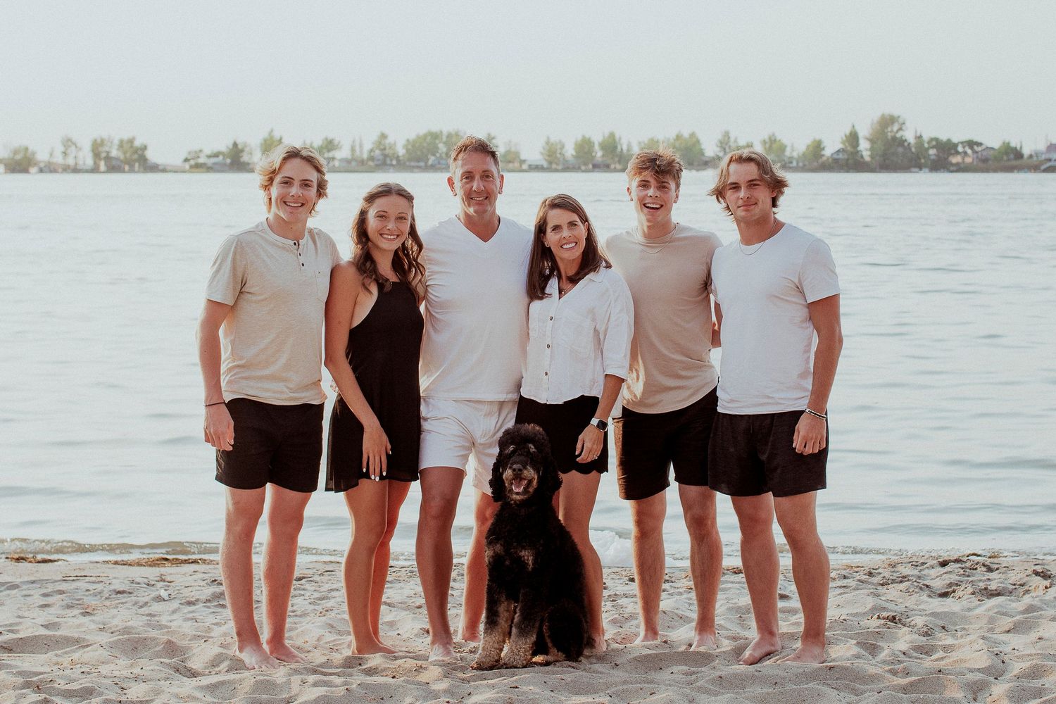 A group in casual summer attire stands together with their black dog on a beach with water and trees in the background.