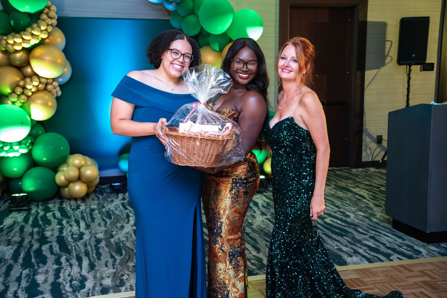 Three people in sparkling green formal dresses pose together at an event with green and gold balloon decorations.