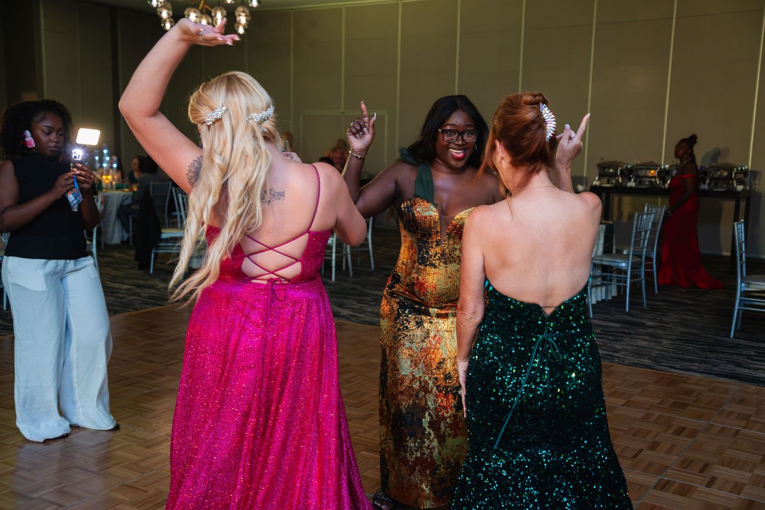 Three people in formal dresses dance together at a reception, wearing pink, floral, and emerald gowns.