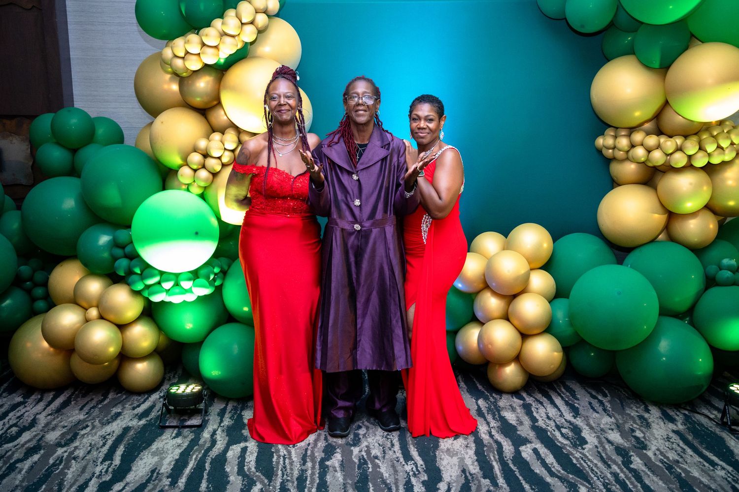 Balloon arch with alternating green and gold balloons against a blue wall with people posing in red and purple outfits.