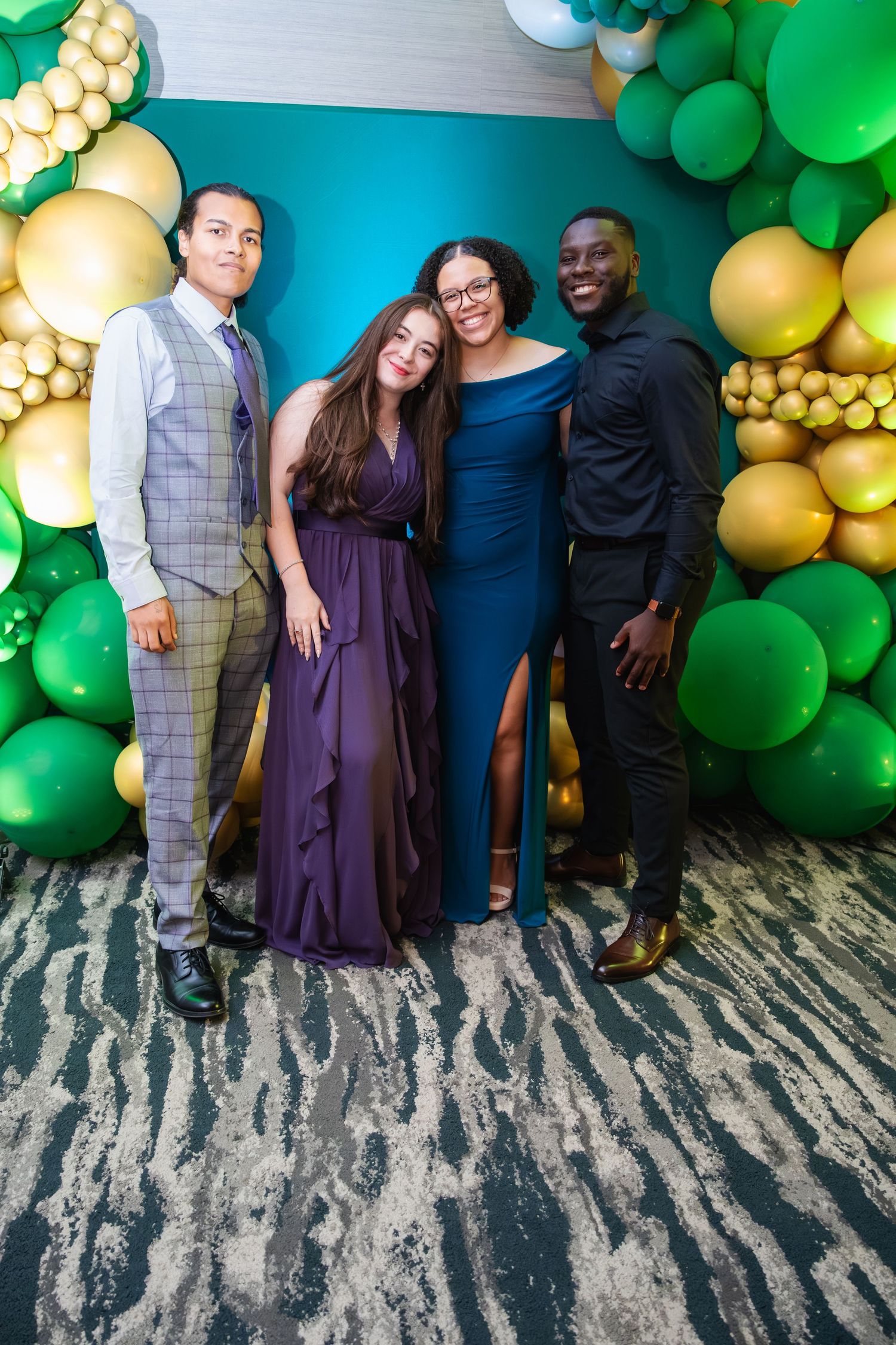 Group poses against decorative wall with green and gold balloon arch display at celebration event.