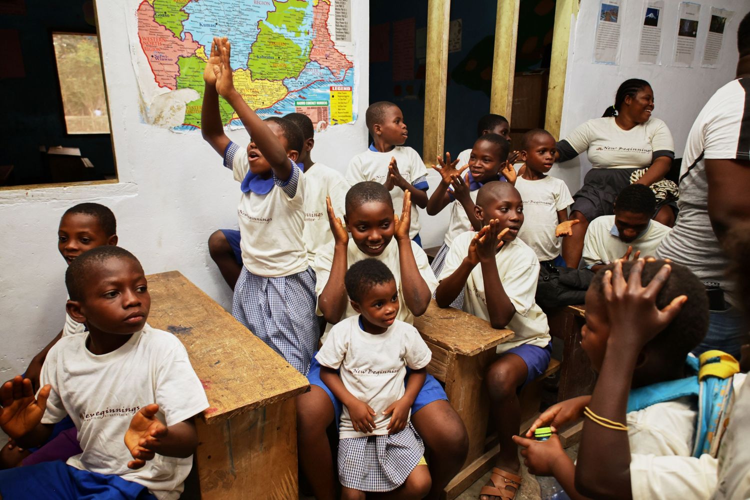 Students in a classroom raise their hands enthusiastically while sitting at wooden desks with a colorful map on the wall.