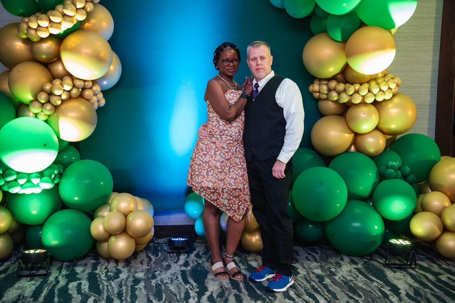 Festive party backdrop with green and gold balloon arch decorations against a blue wall.