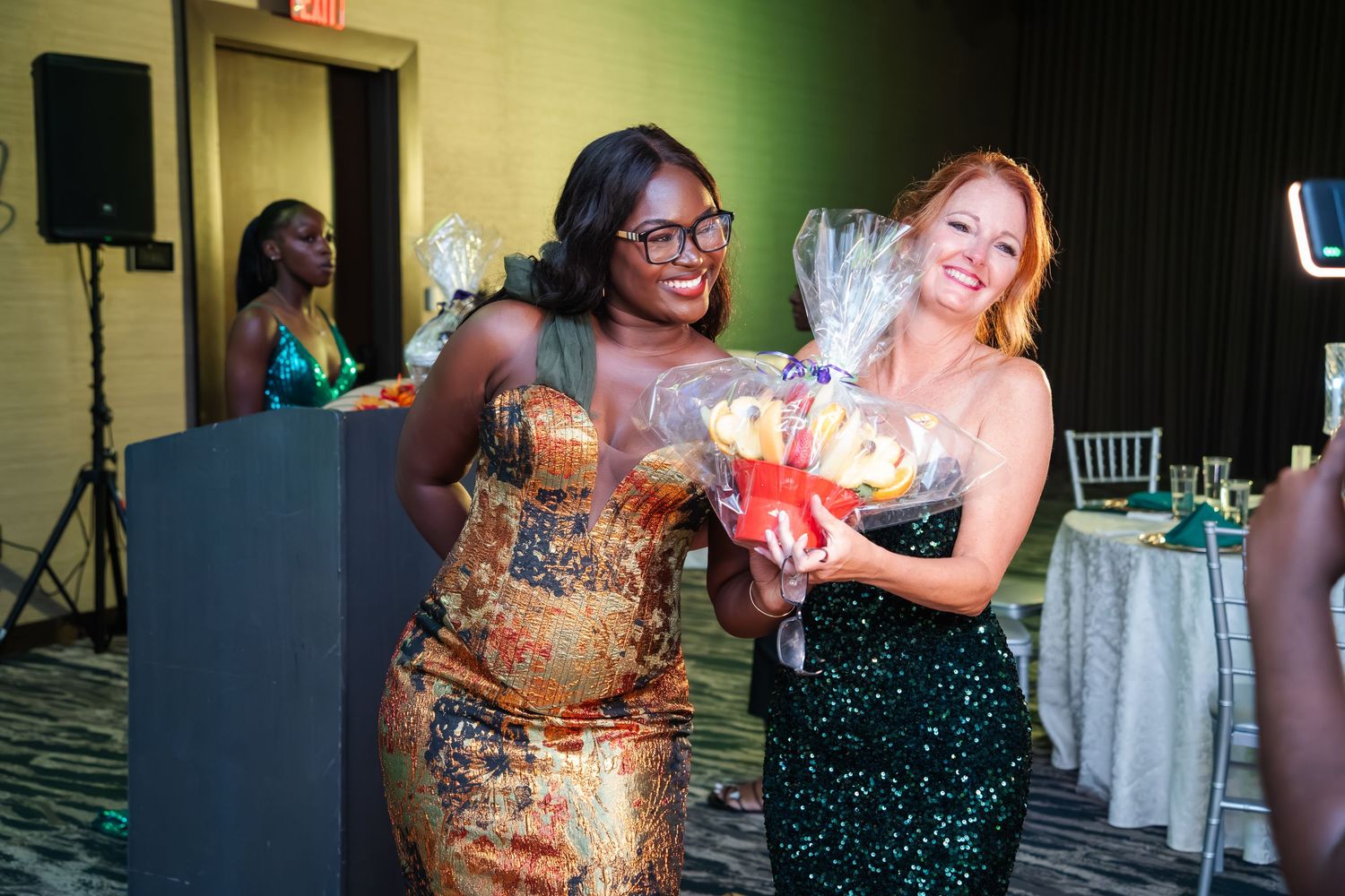 Two people in sparkly evening dresses laugh and celebrate together at a festive indoor event.