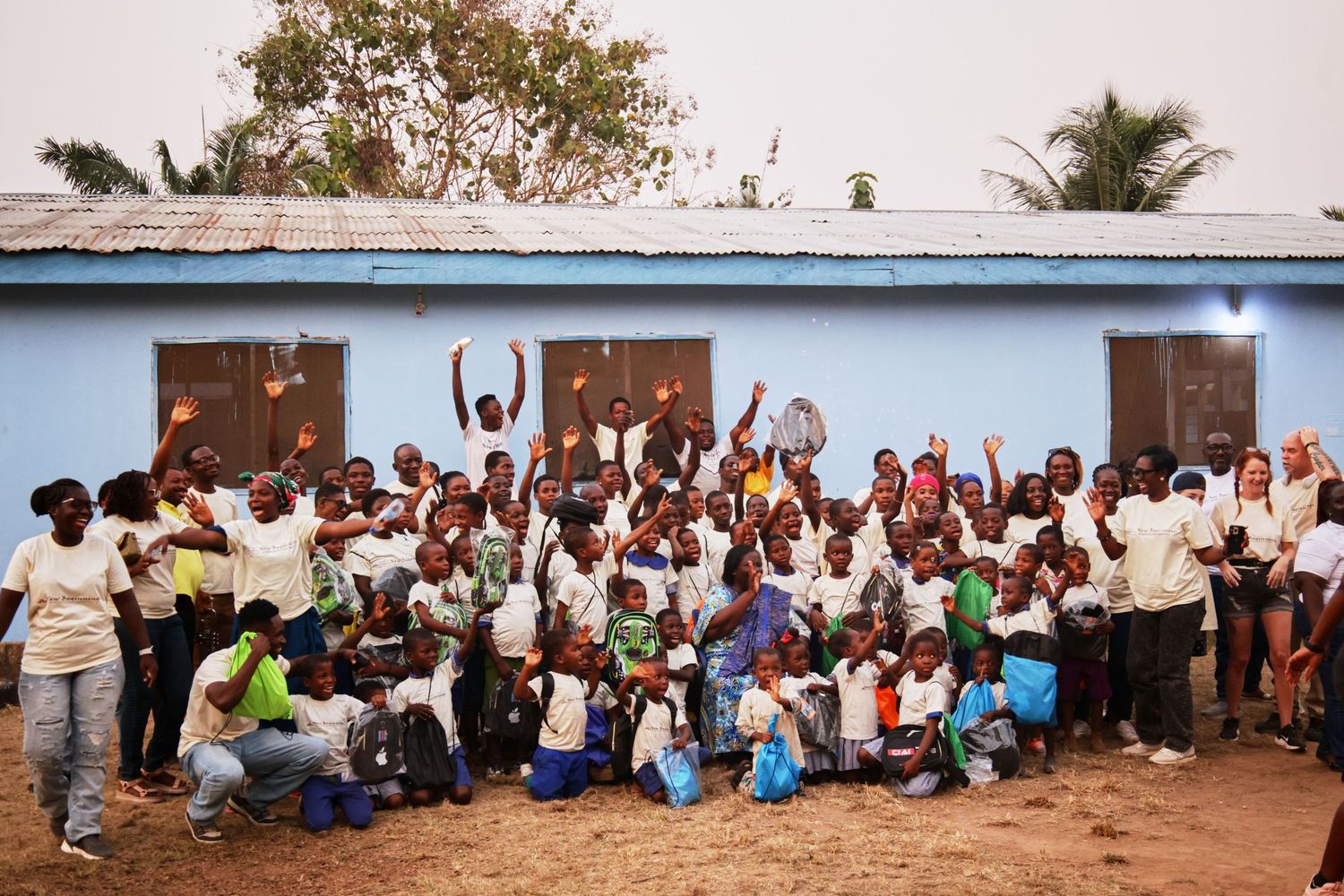 Large group poses cheerfully outside a blue building with raised hands and bright smiles.