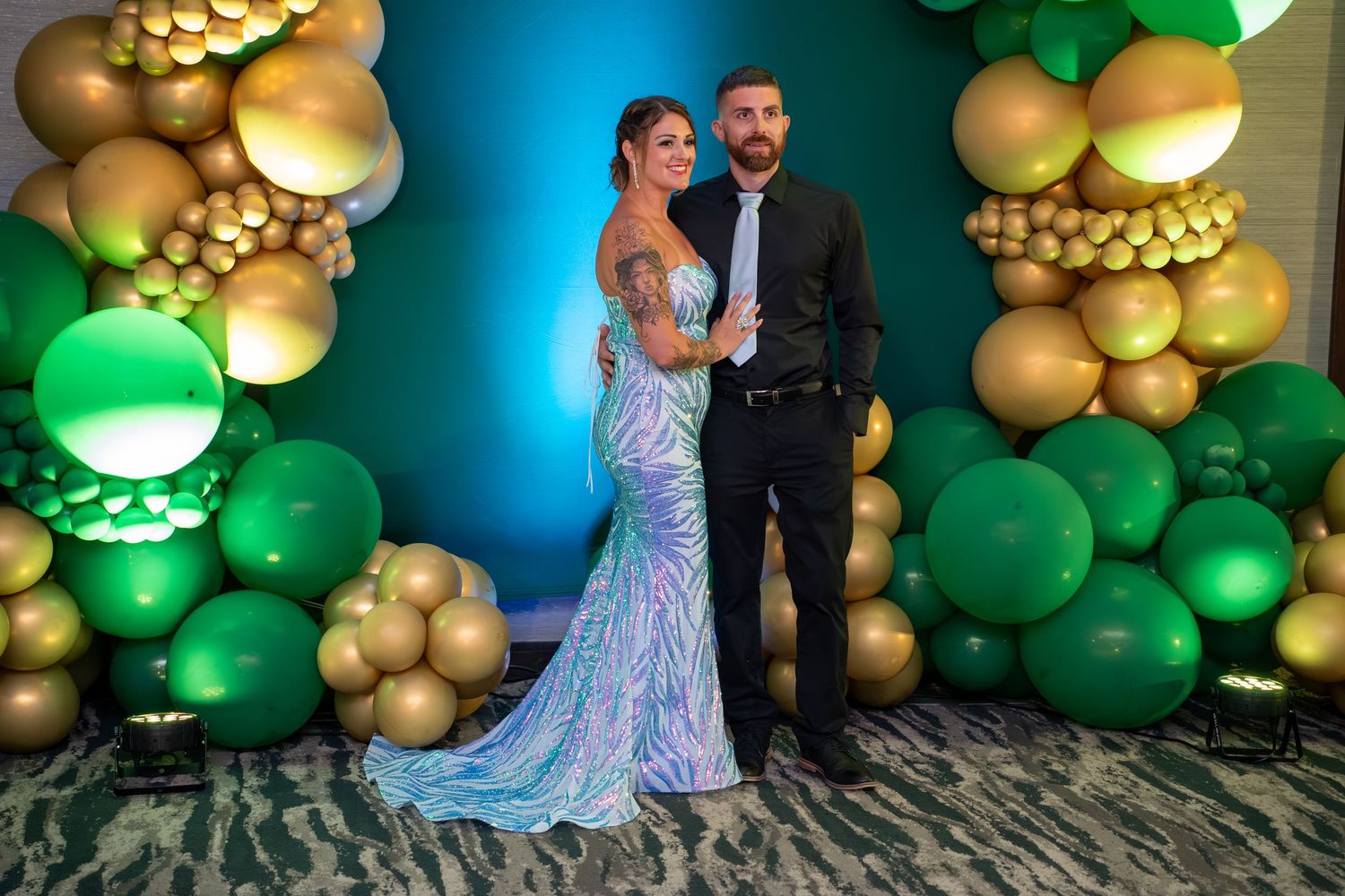 A couple celebrates at a wedding reception with green and gold balloon decorations against a blue-lit backdrop.