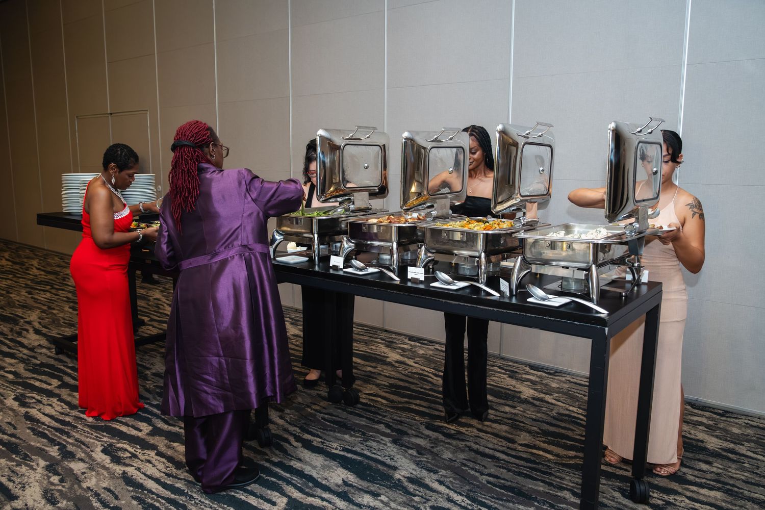 Guests in formal attire serve themselves at an elegant buffet line with multiple chafing dishes on a dark patterned carpet.