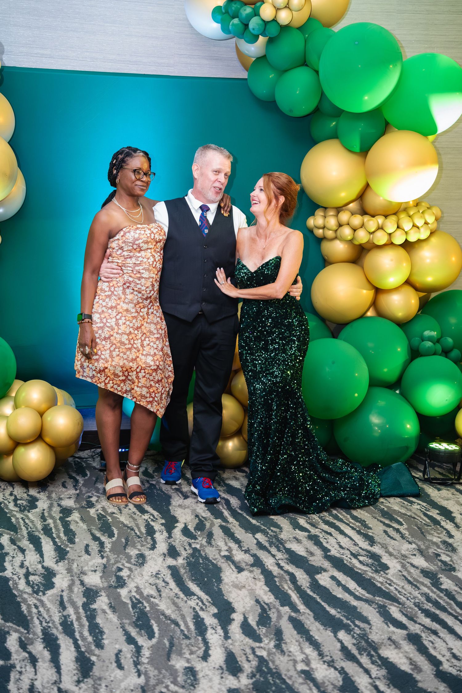 Three people pose together in front of a green and gold balloon arch backdrop at a formal event.