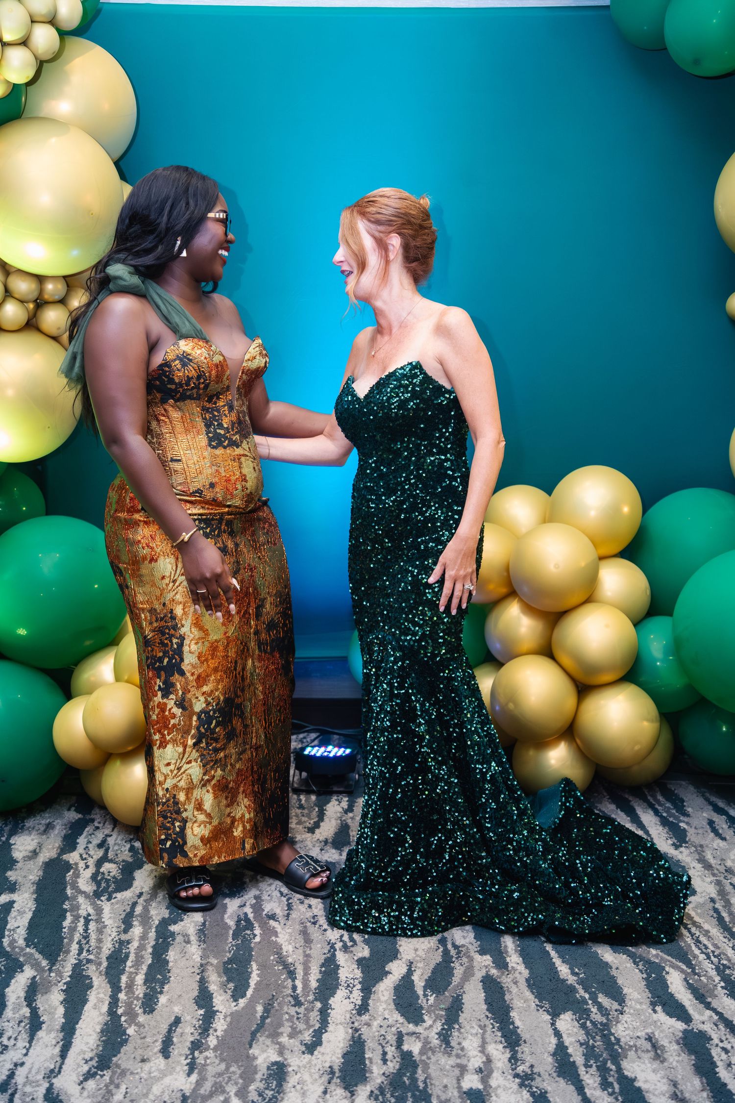 Two people in elegant evening gowns, one gold and one emerald green, pose against a backdrop of gold and green balloons.