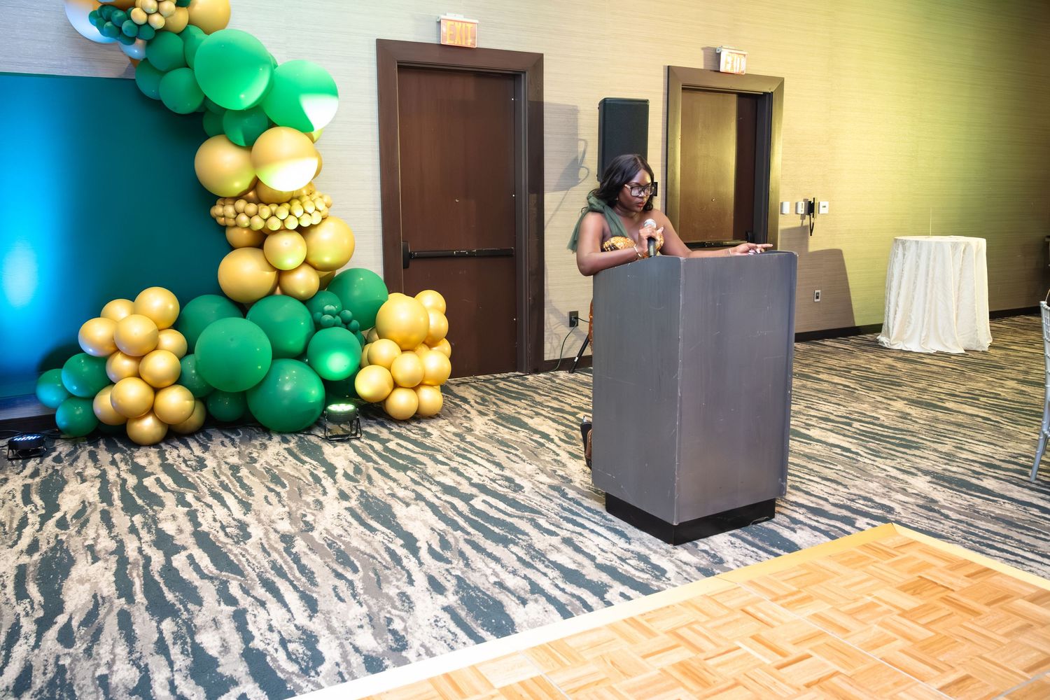 Elegant balloon arch installation with green and gold balloons decorating an indoor venue hallway.