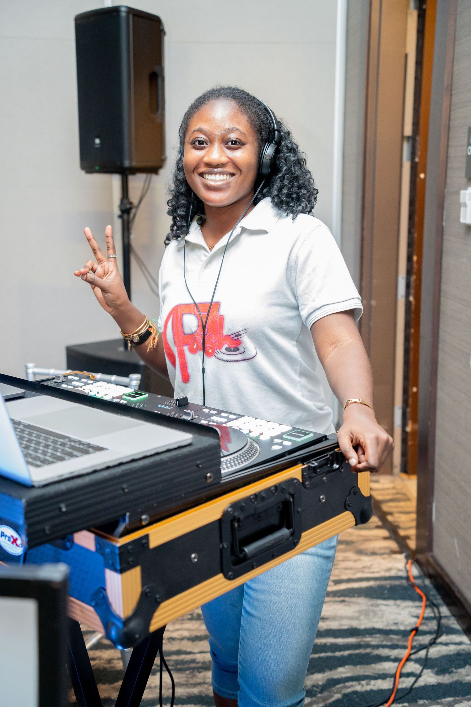 DJ stands at mixing equipment with headphones and a white polo shirt, making a peace sign while smiling.