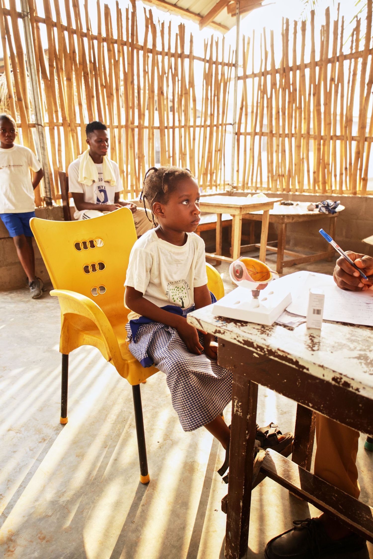 Student sits at wooden desk in classroom with bamboo walls and sunlight streaming through.