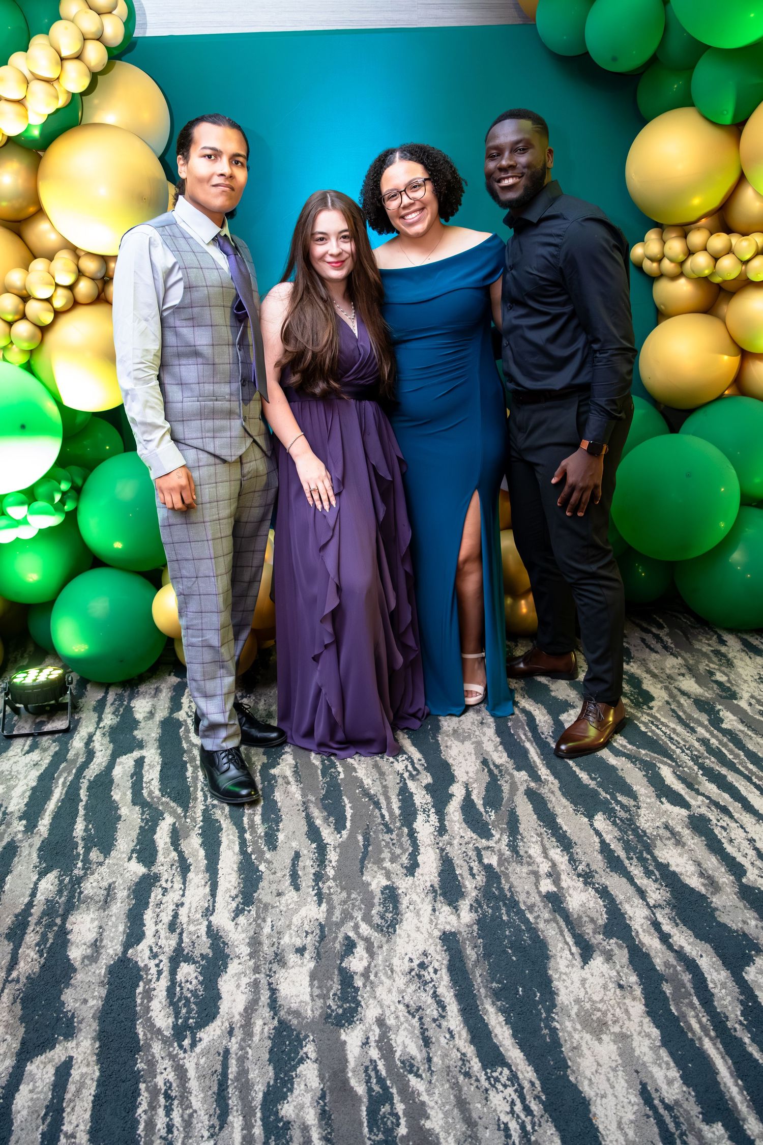 A group poses in front of a festive balloon arch with green and gold balloons against a marbled floor.